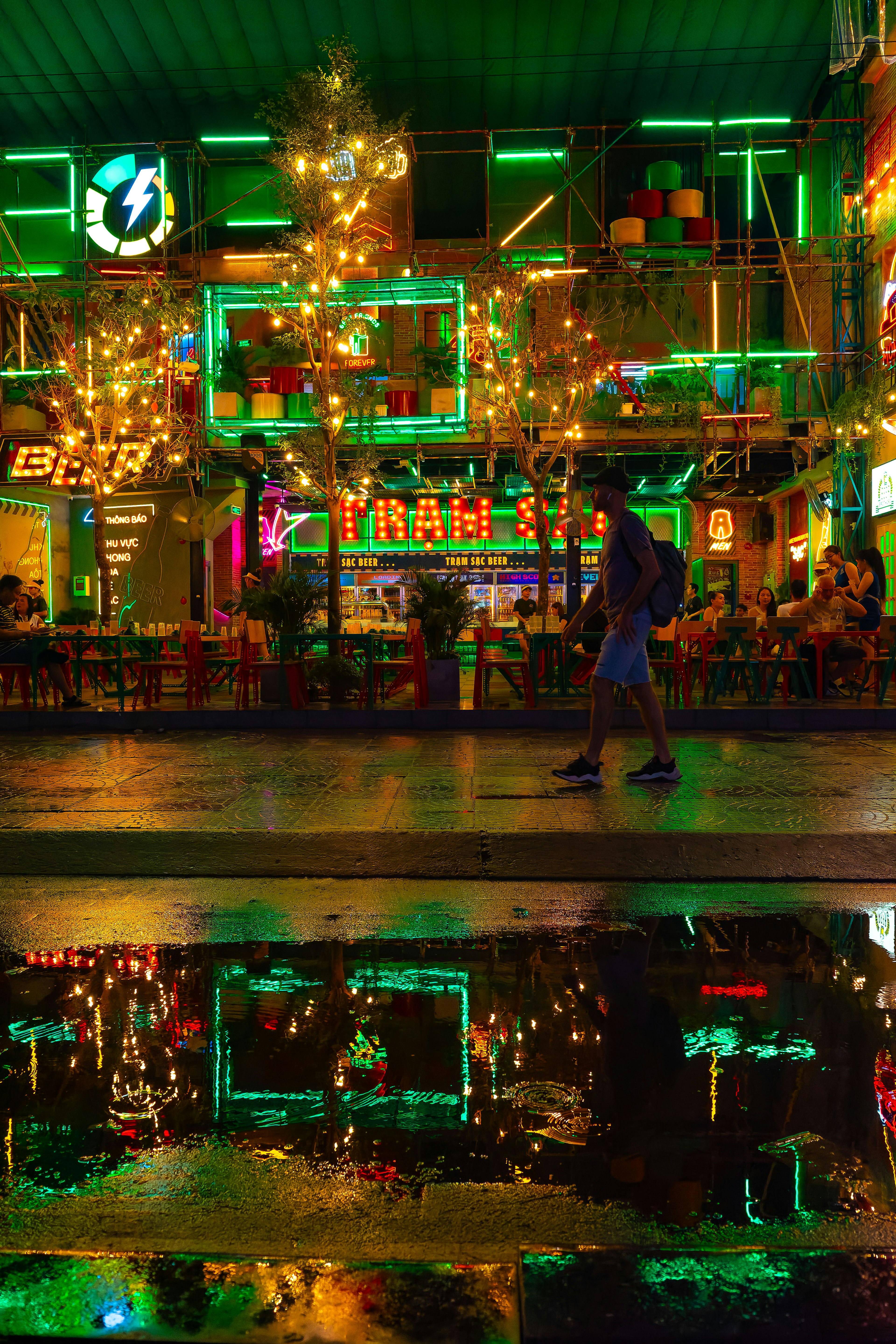 Colorful neon lights illuminate an outdoor bar scene with a person walking