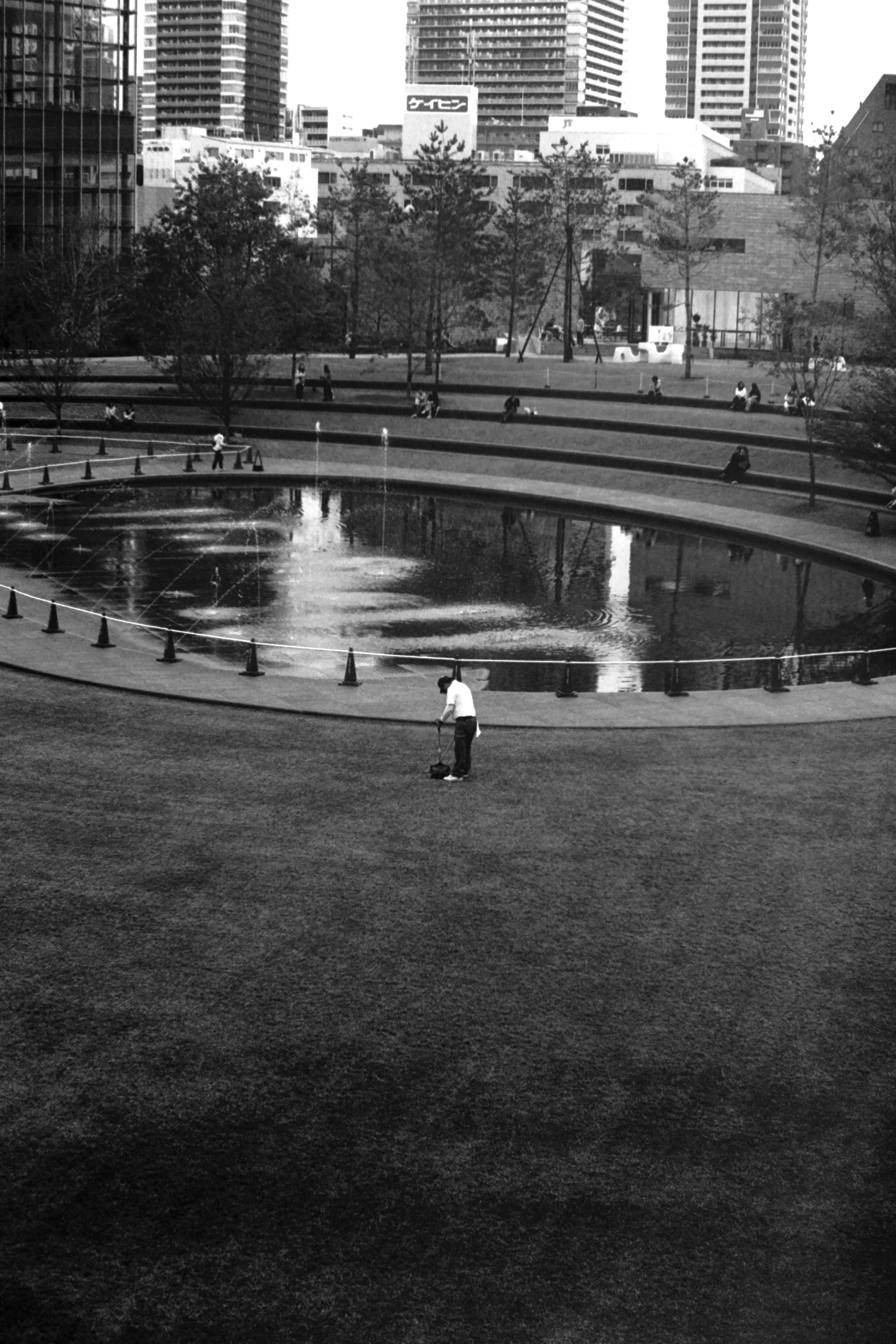 Person standing in a green area near a circular pond surrounded by city buildings