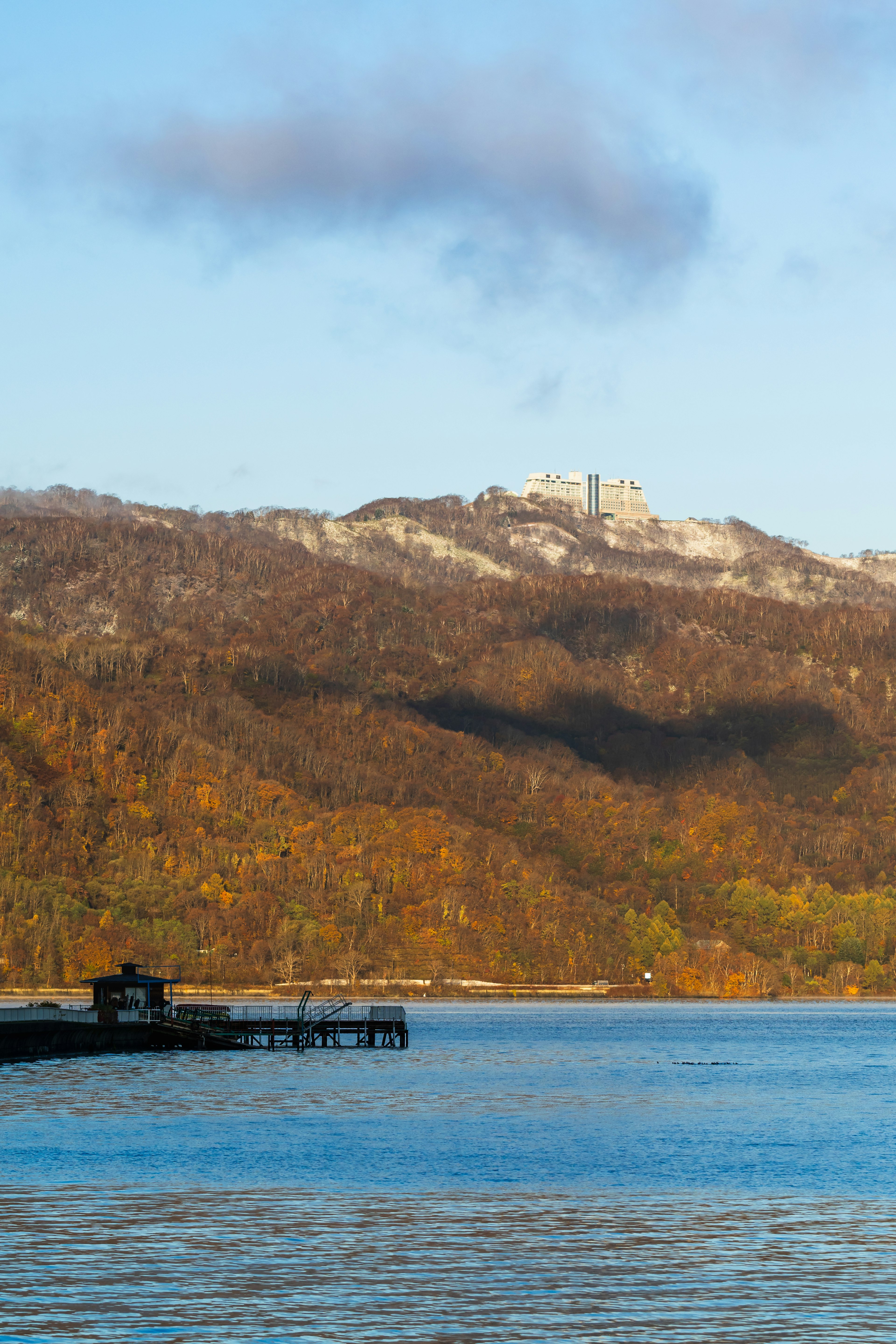 Ruhiger See mit bunten Bergen im Hintergrund und einem nahegelegenen Pier und Gebäude