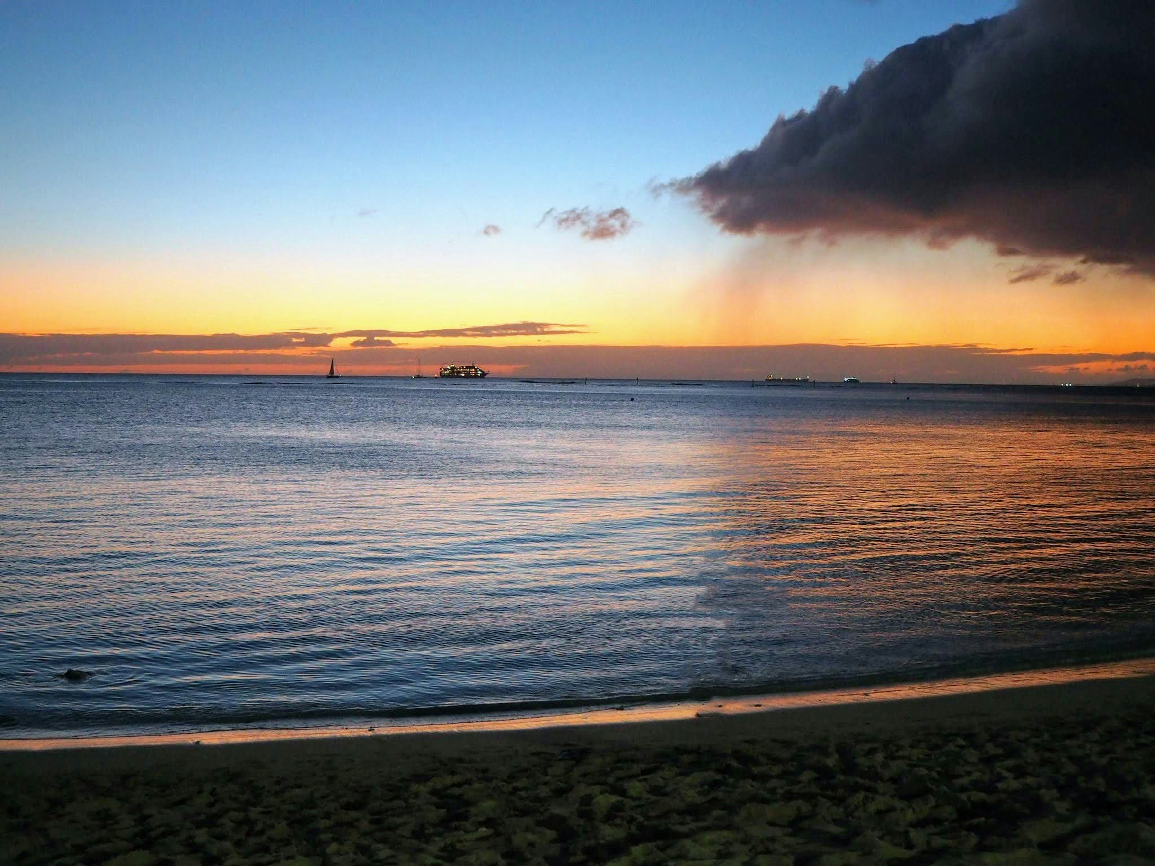 Atardecer sobre el océano con una playa, olas tranquilas y barcos a lo lejos