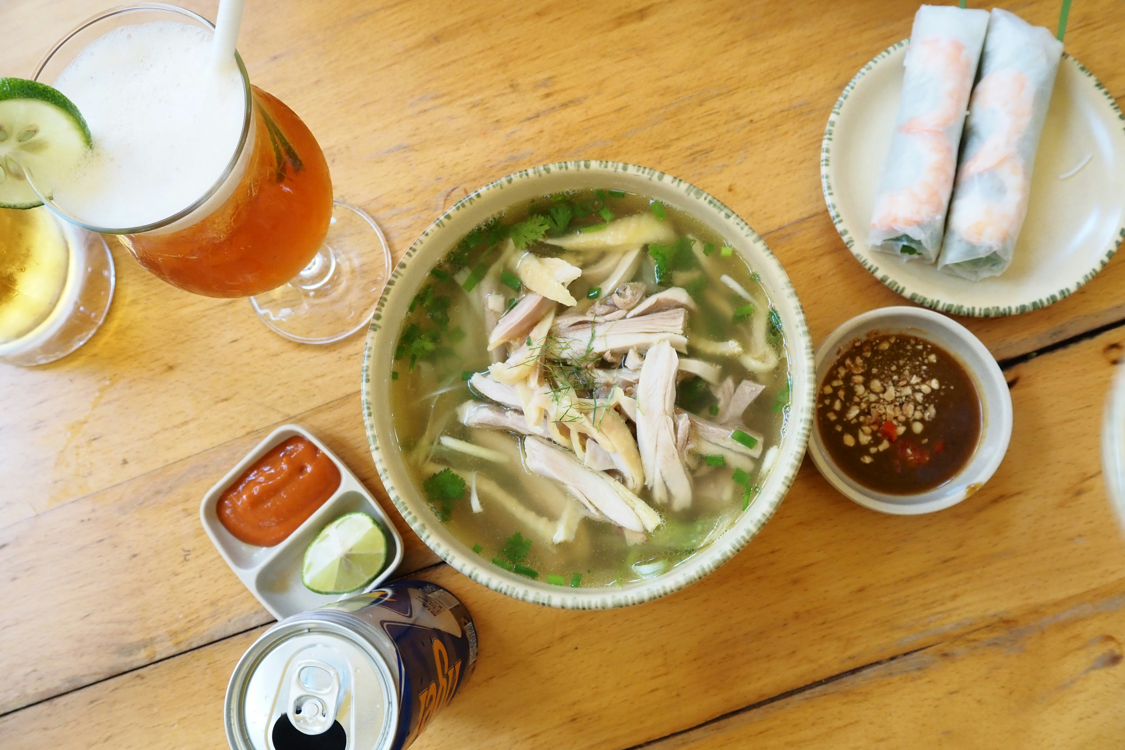 A bowl of pho with chicken and herbs accompanied by spring rolls and drinks on a wooden table