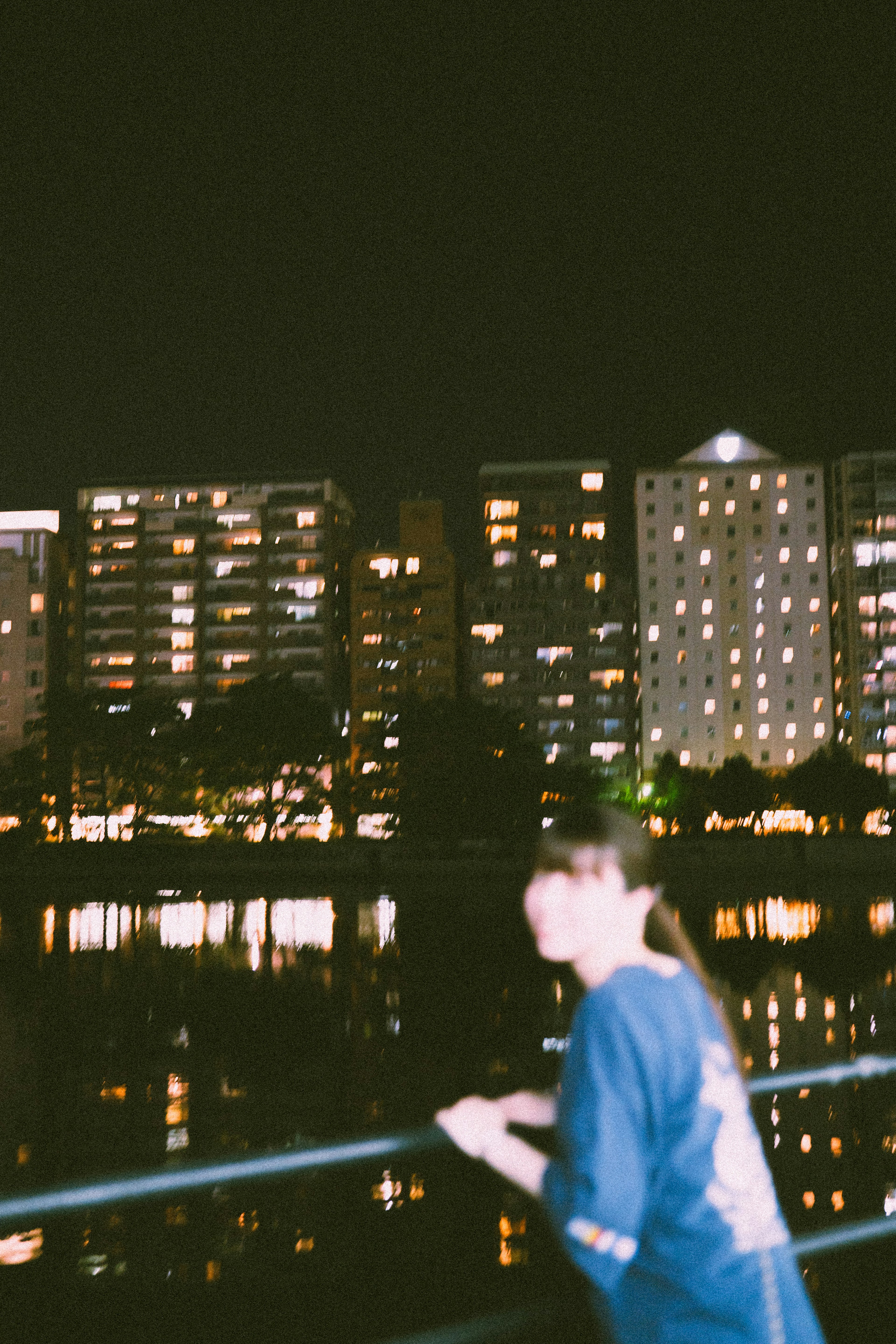 Una persona apoyada en una barandilla con luces de la ciudad y reflejos en el agua de noche