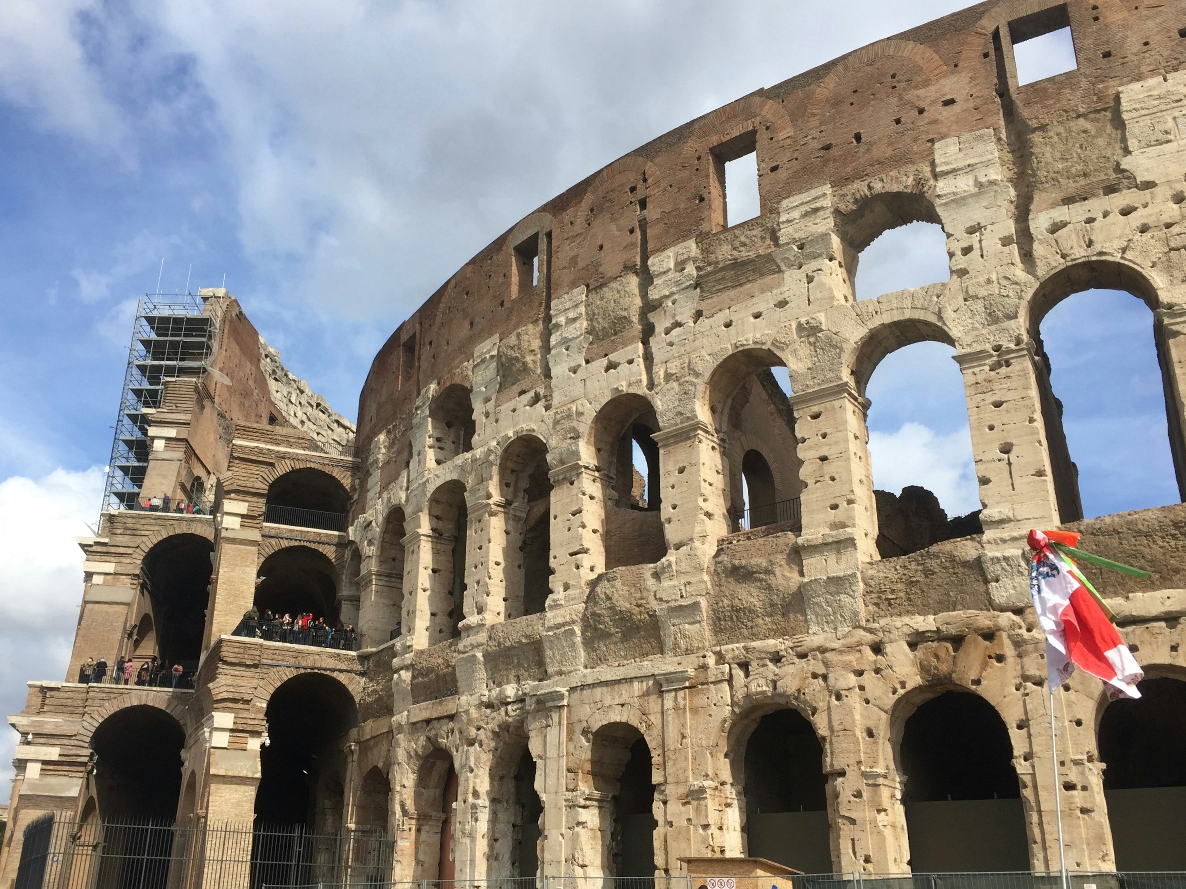 Vista esterna del Colosseo in restauro, cielo blu con nuvole, bandiera italiana