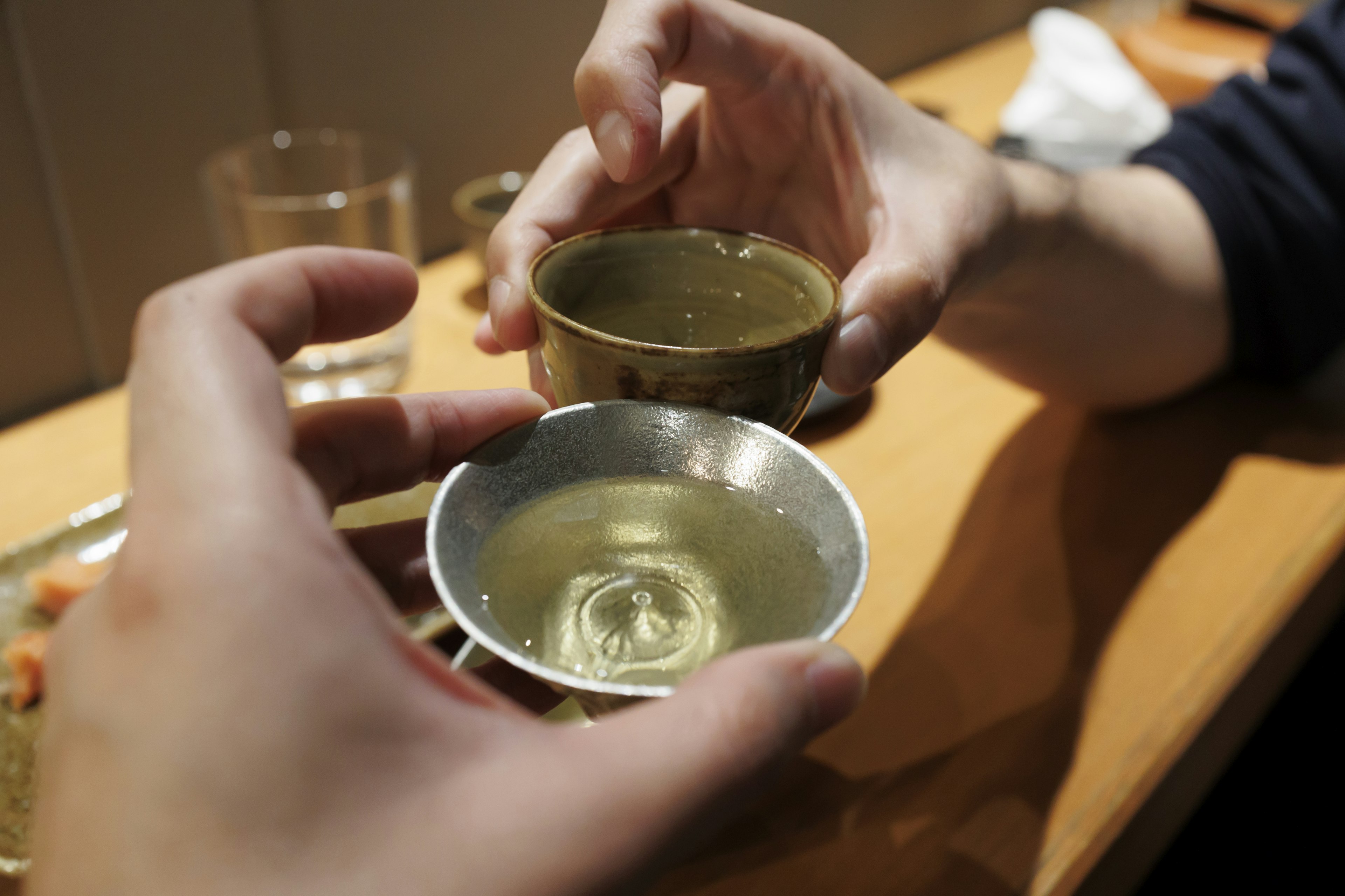 Manos sosteniendo dos copas de sake cruzándose sobre una mesa con sake