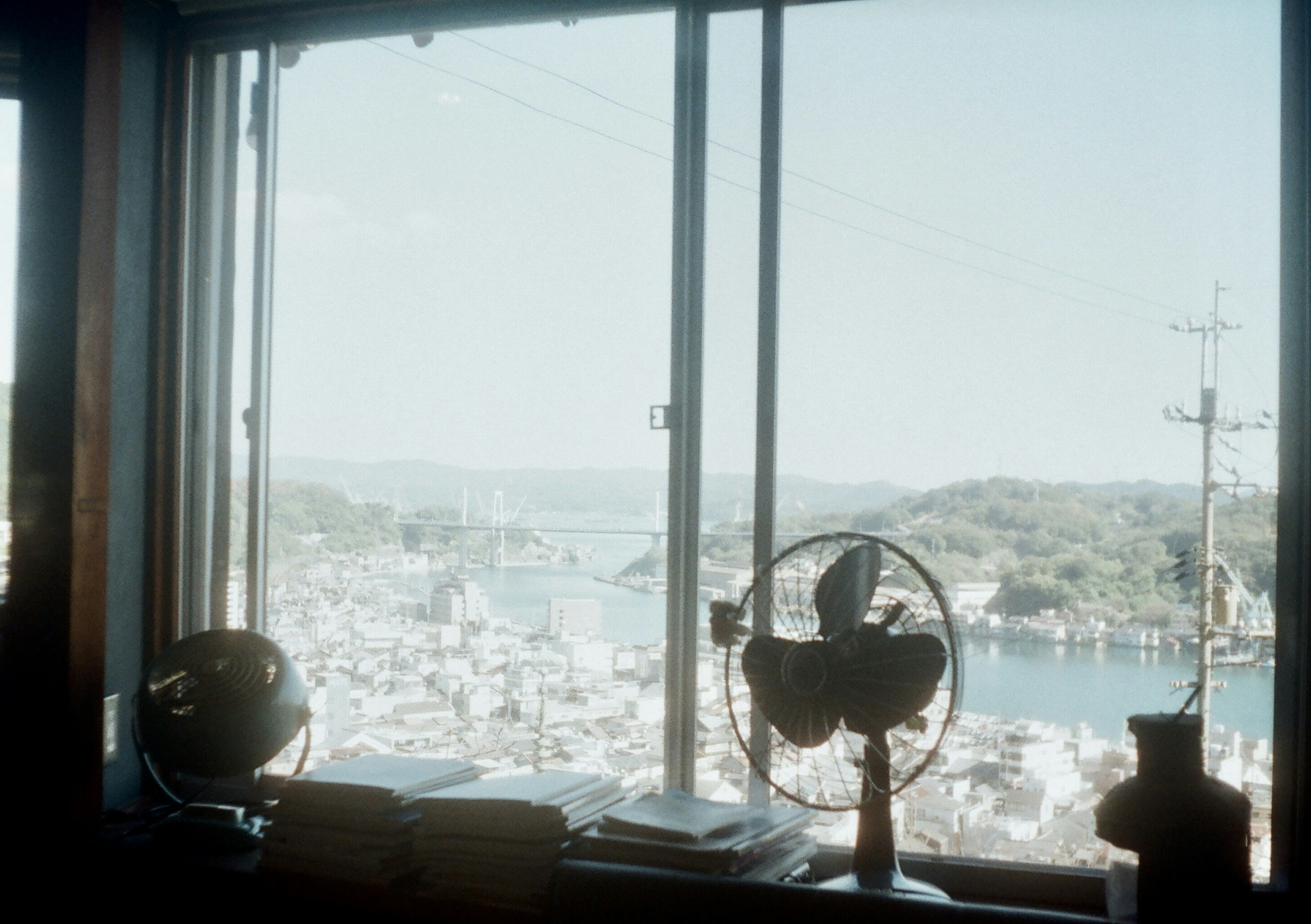 Vista interior de una habitación con un ventilador antiguo y libros con vista a un puerto pintoresco