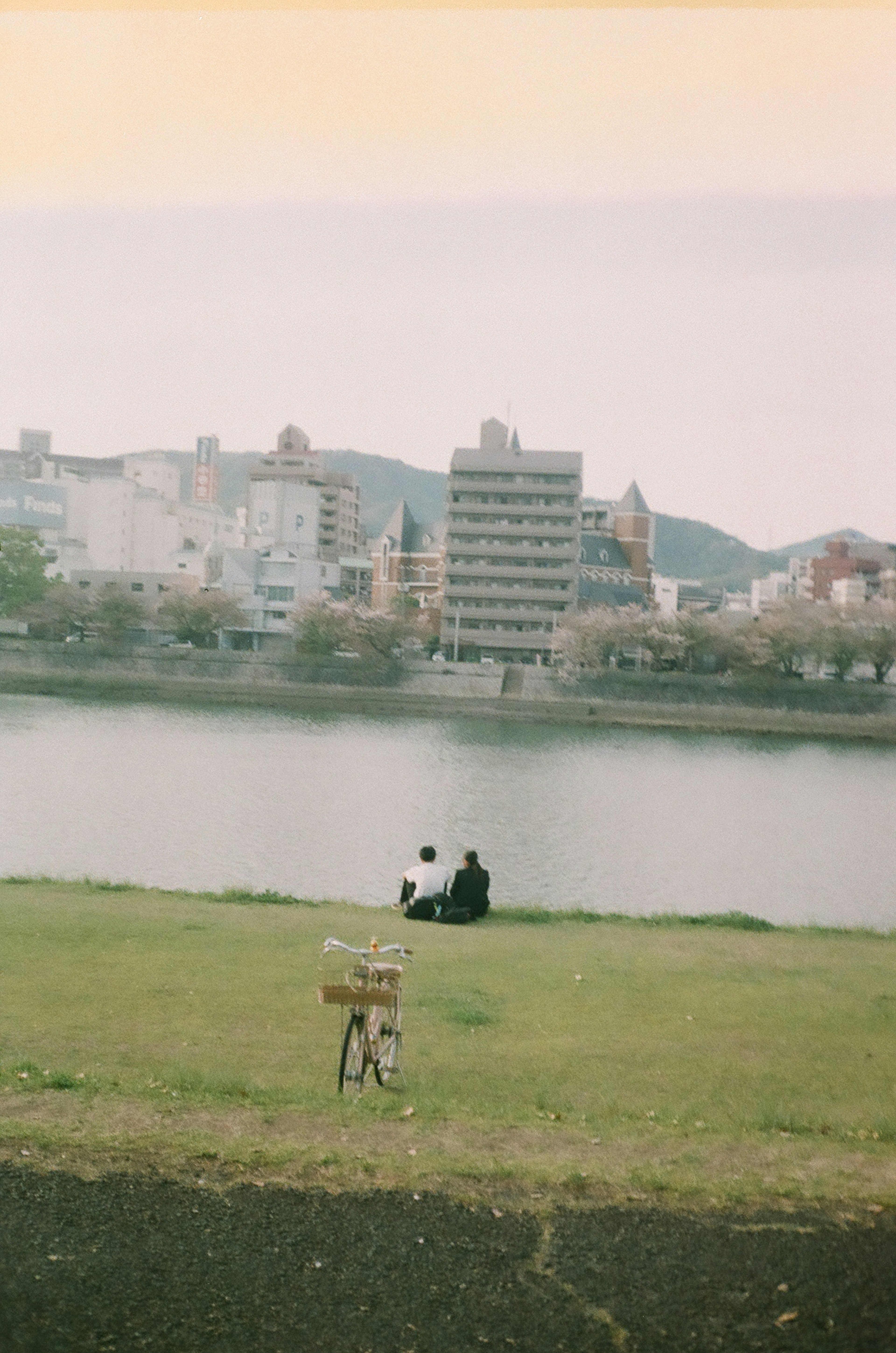 Silhouette of two people sitting by a riverbank in a park with a soft color palette