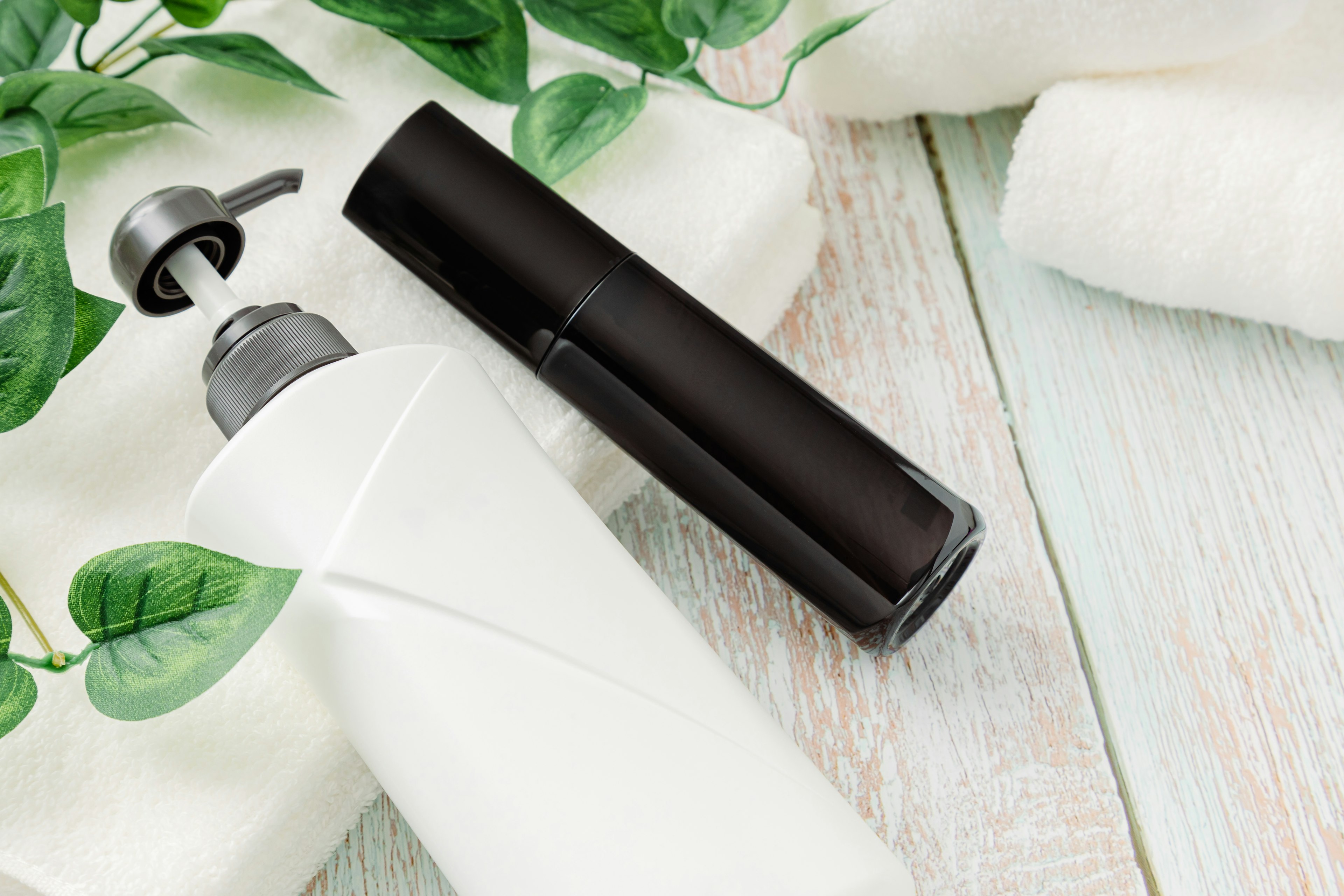 Image of a white bottle and a black bottle of cosmetic products with green leaves and white towels in the background