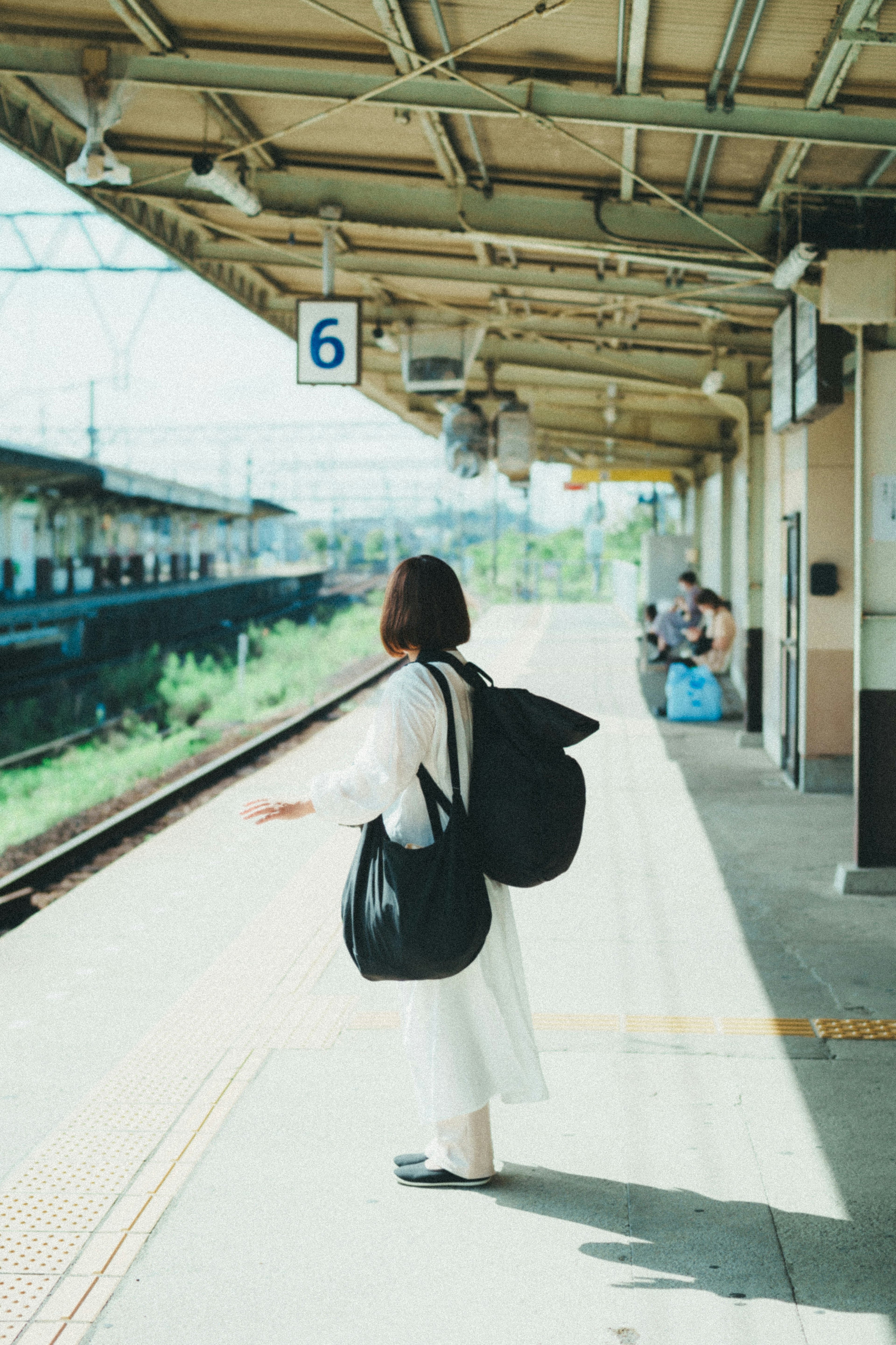 Una mujer de pie en una plataforma de tren vestida de blanco y llevando una bolsa grande