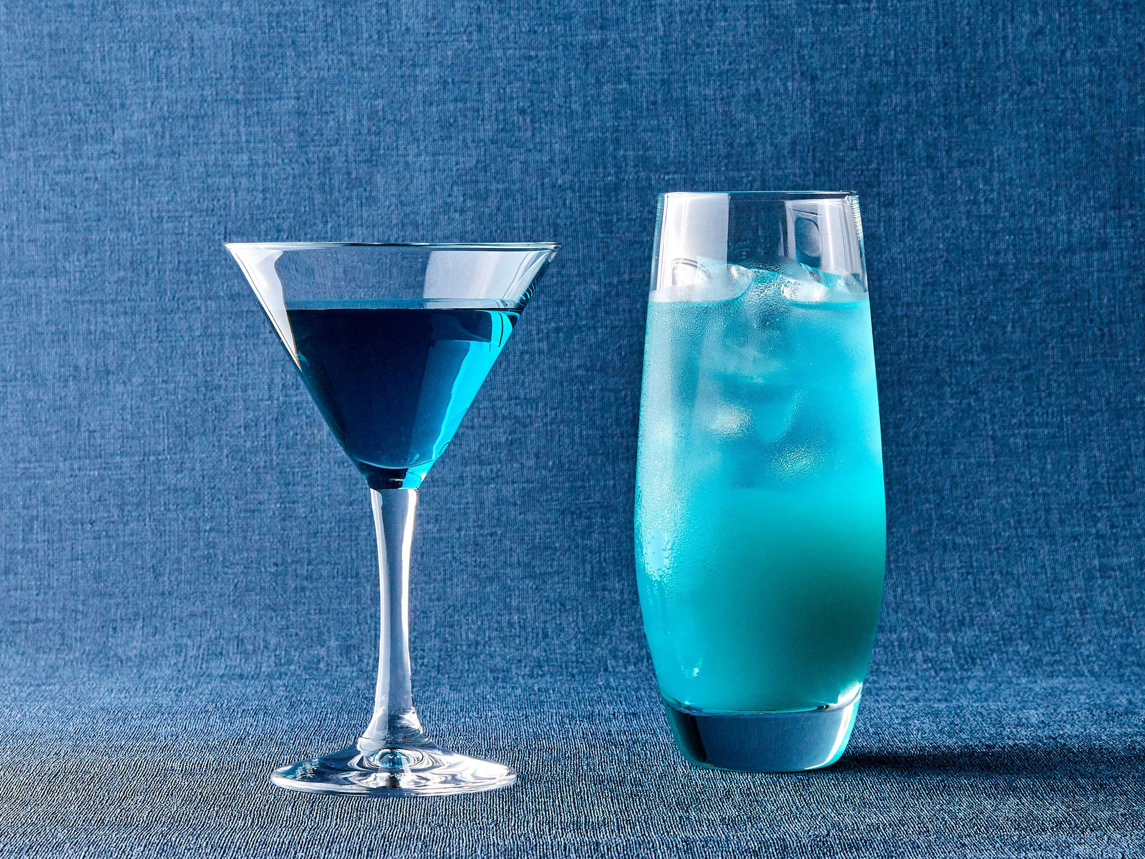 Two blue drinks a martini glass and a tall glass with ice against a blue background