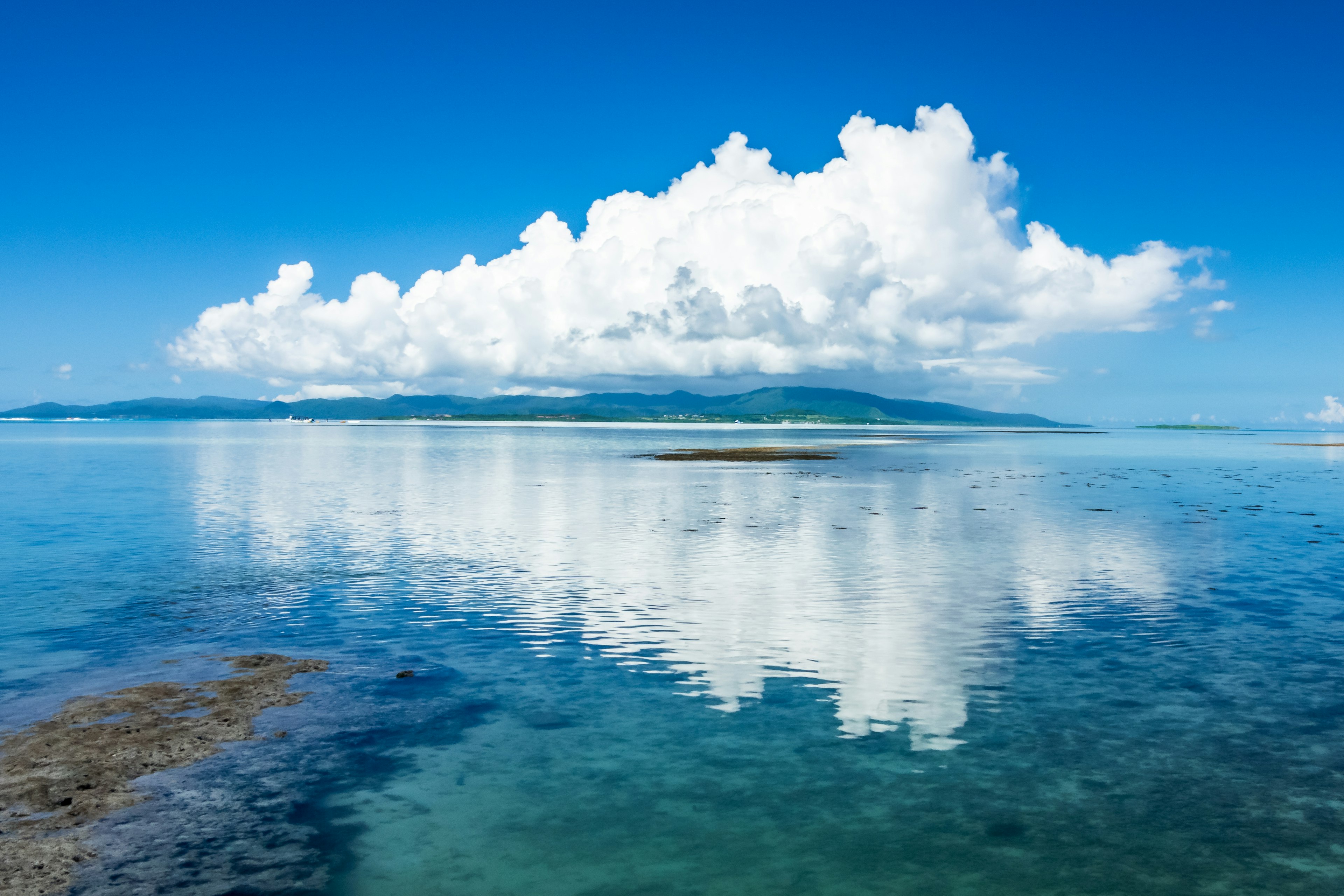 Pemandangan laut tenang dengan langit biru dan awan putih yang tercermin di permukaan air tenang