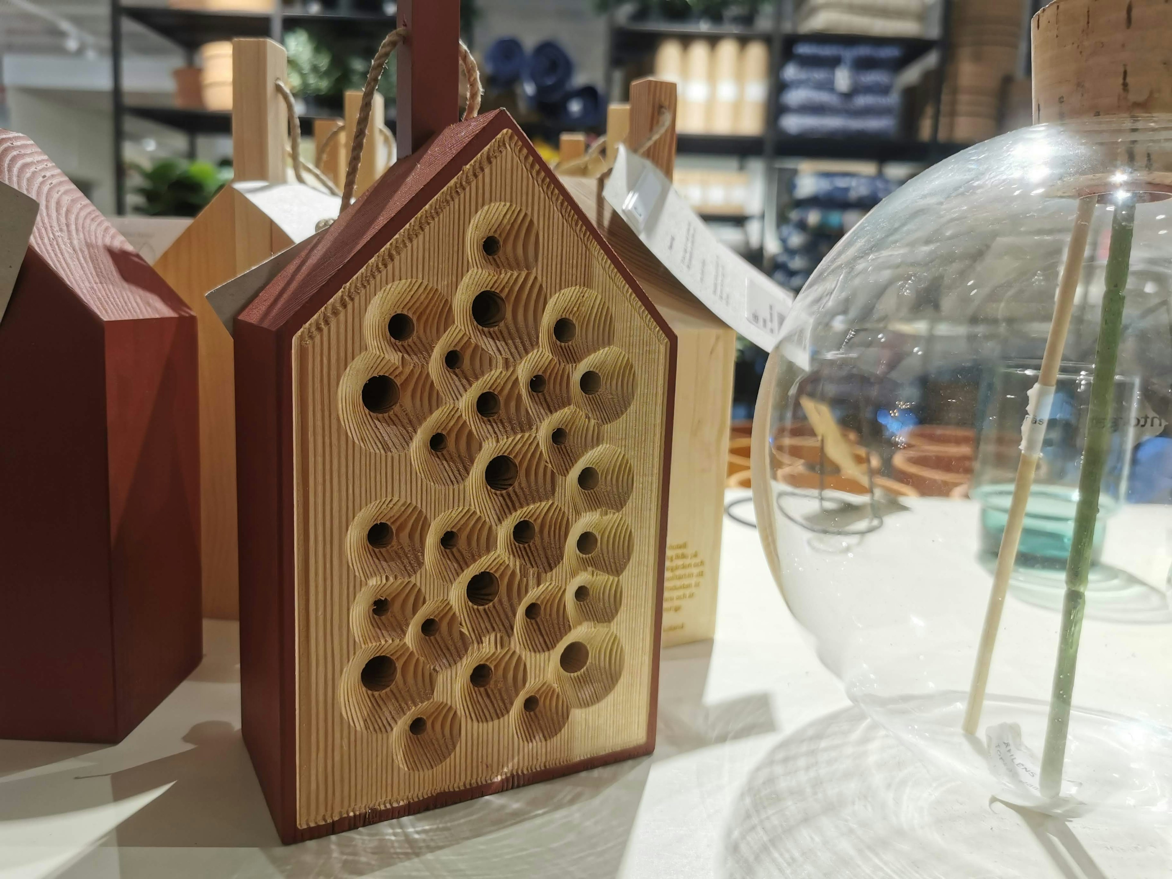 Wooden house-shaped insect hotel with multiple holes beside a transparent glass object