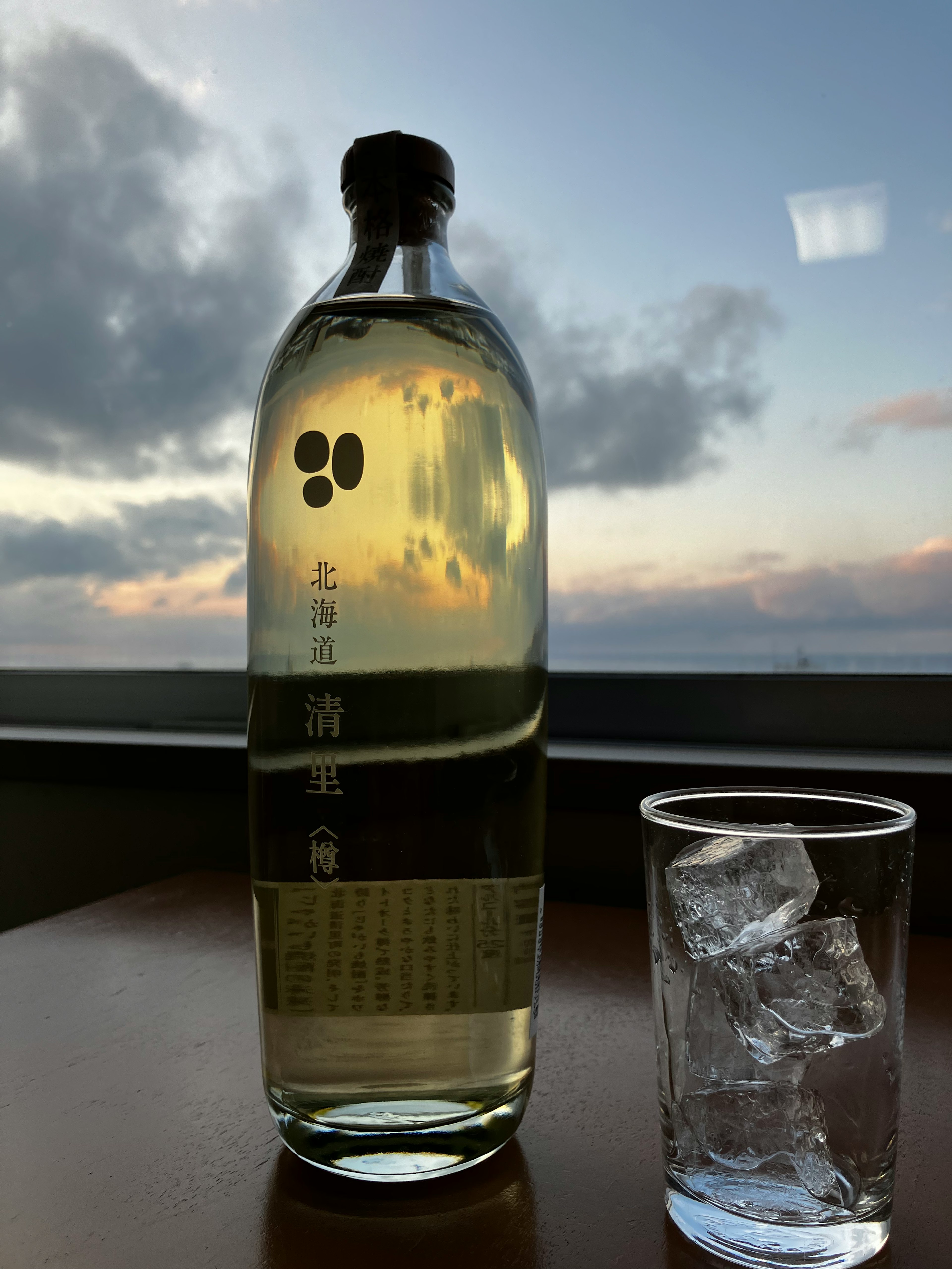 Ein klarer Drink in einer Flasche und ein Glas mit Eis, das am Fenster steht, mit einem schönen Sonnenuntergang im Hintergrund