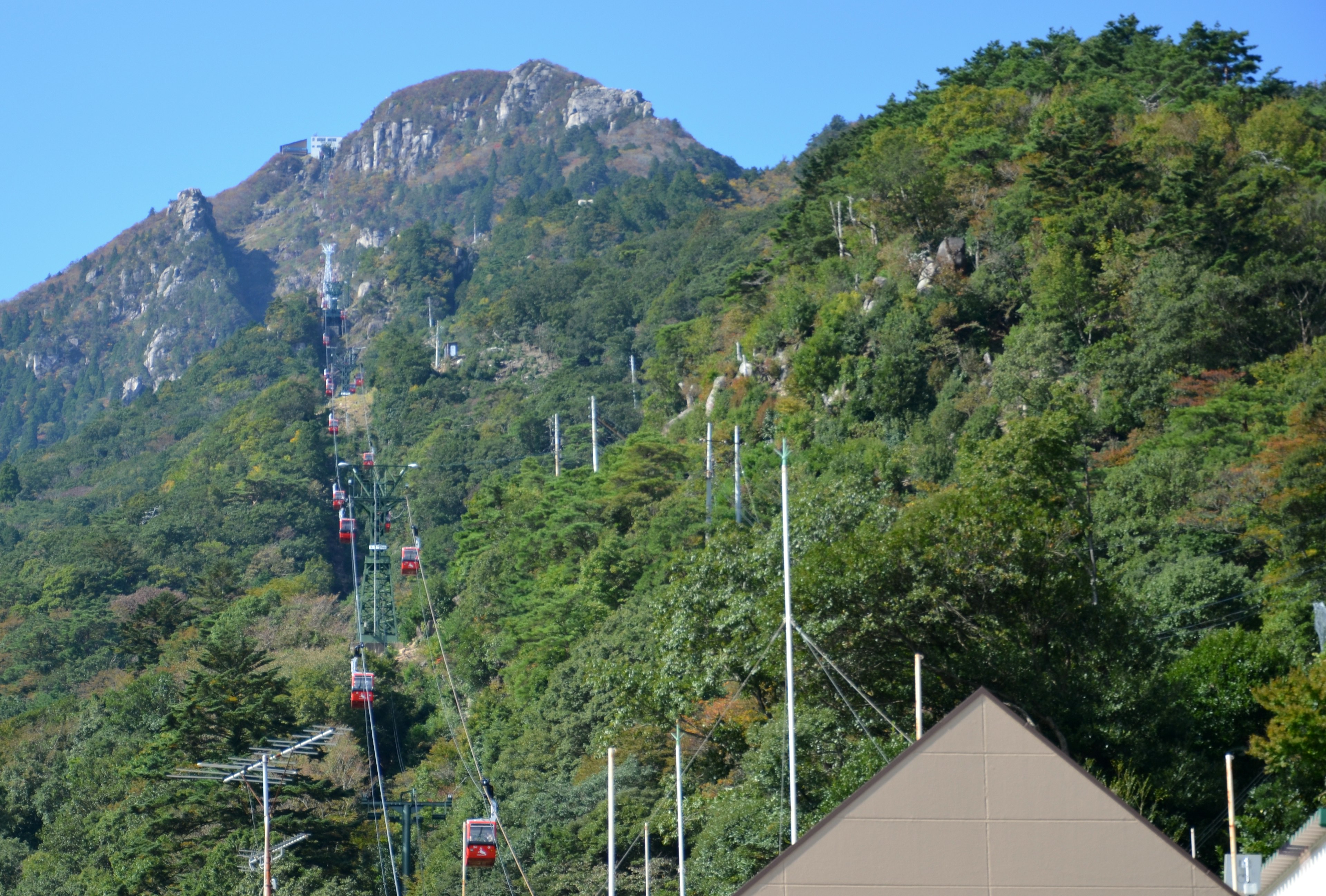 Malersicht auf eine Seilbahn, die einen Berghang mit grünen Bäumen und blauem Himmel hinauffährt