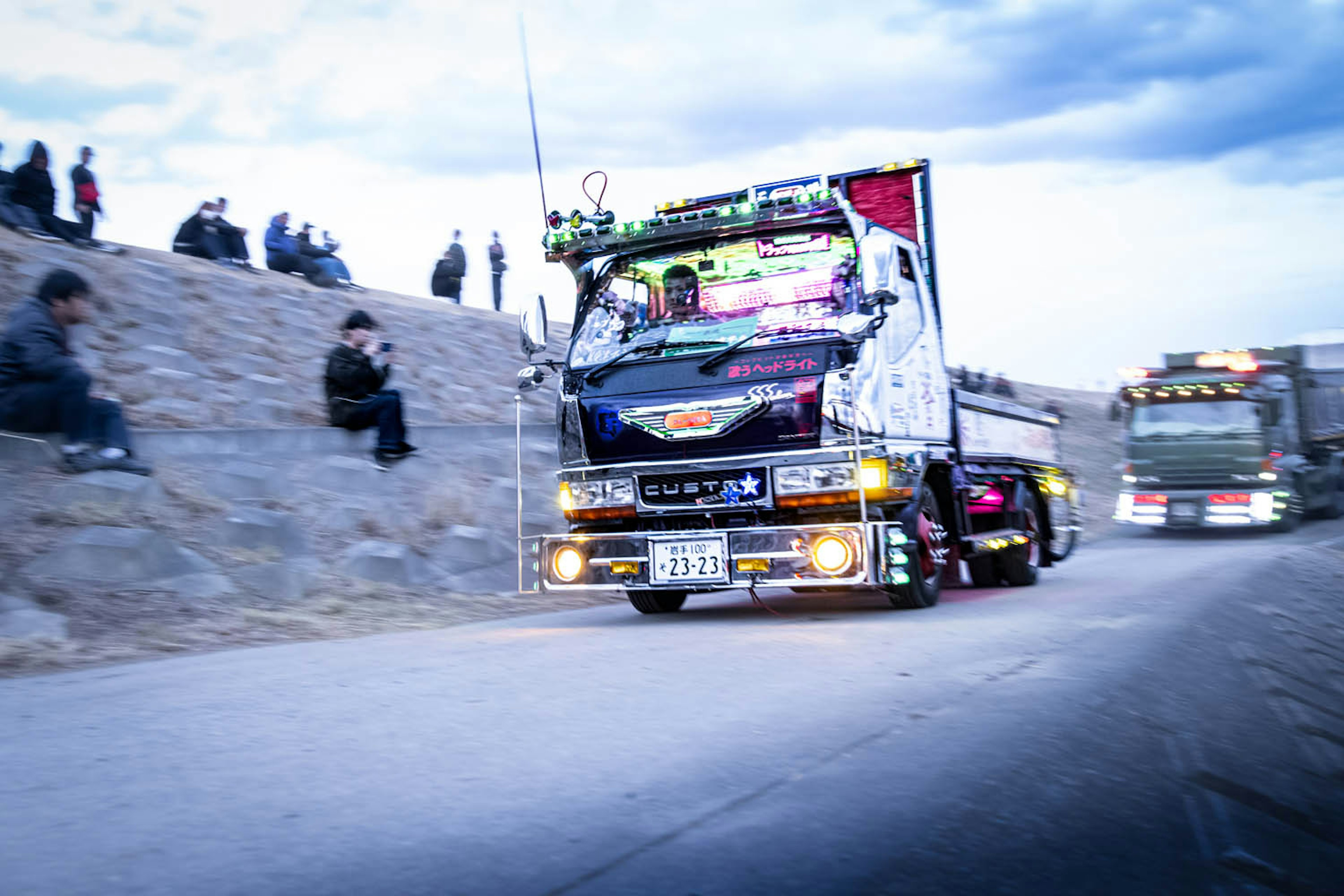 A colorful truck driving on the road with spectators sitting on the side