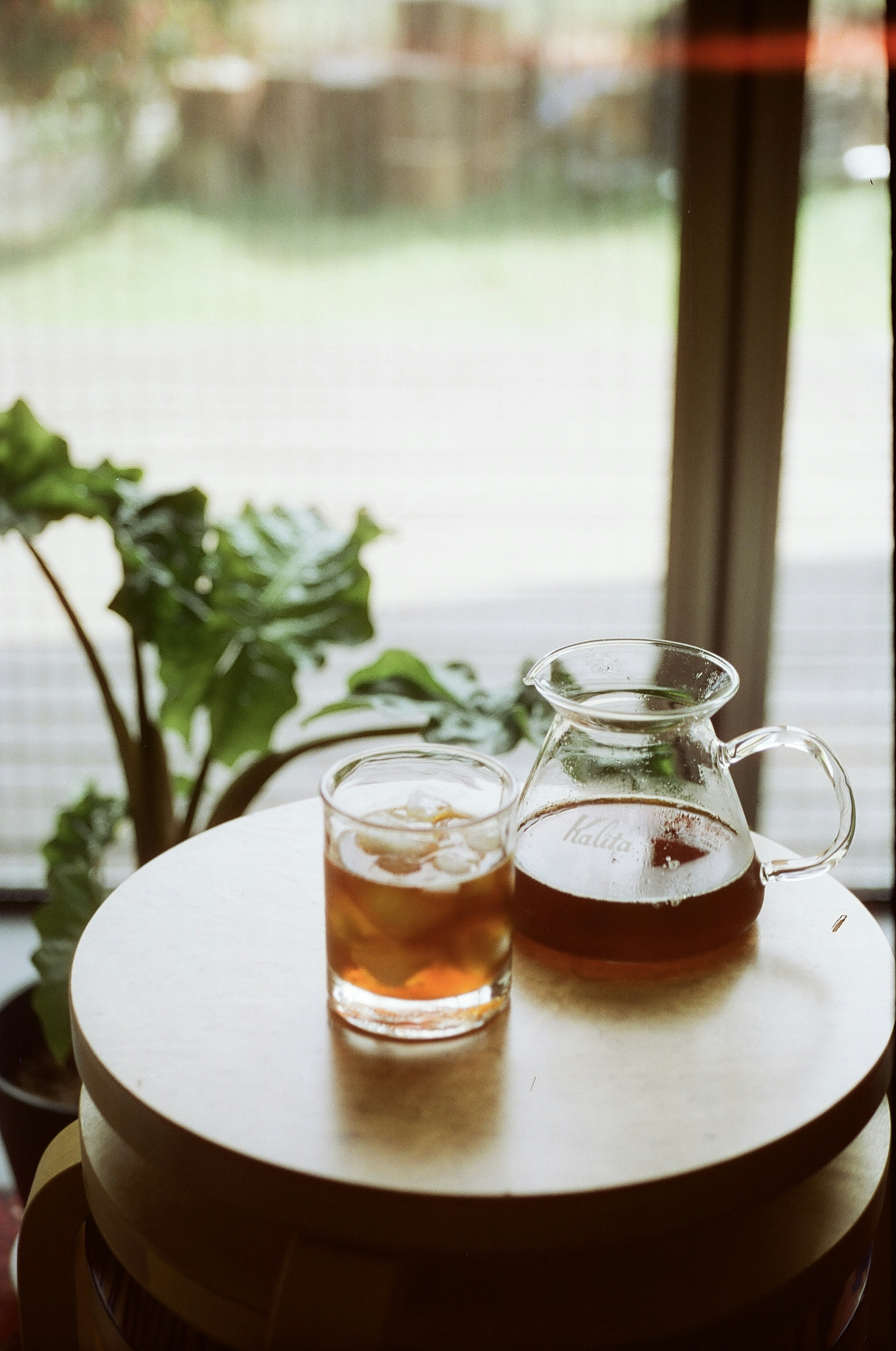 A glass of iced tea and a pot on a wooden table with greenery visible outside the window