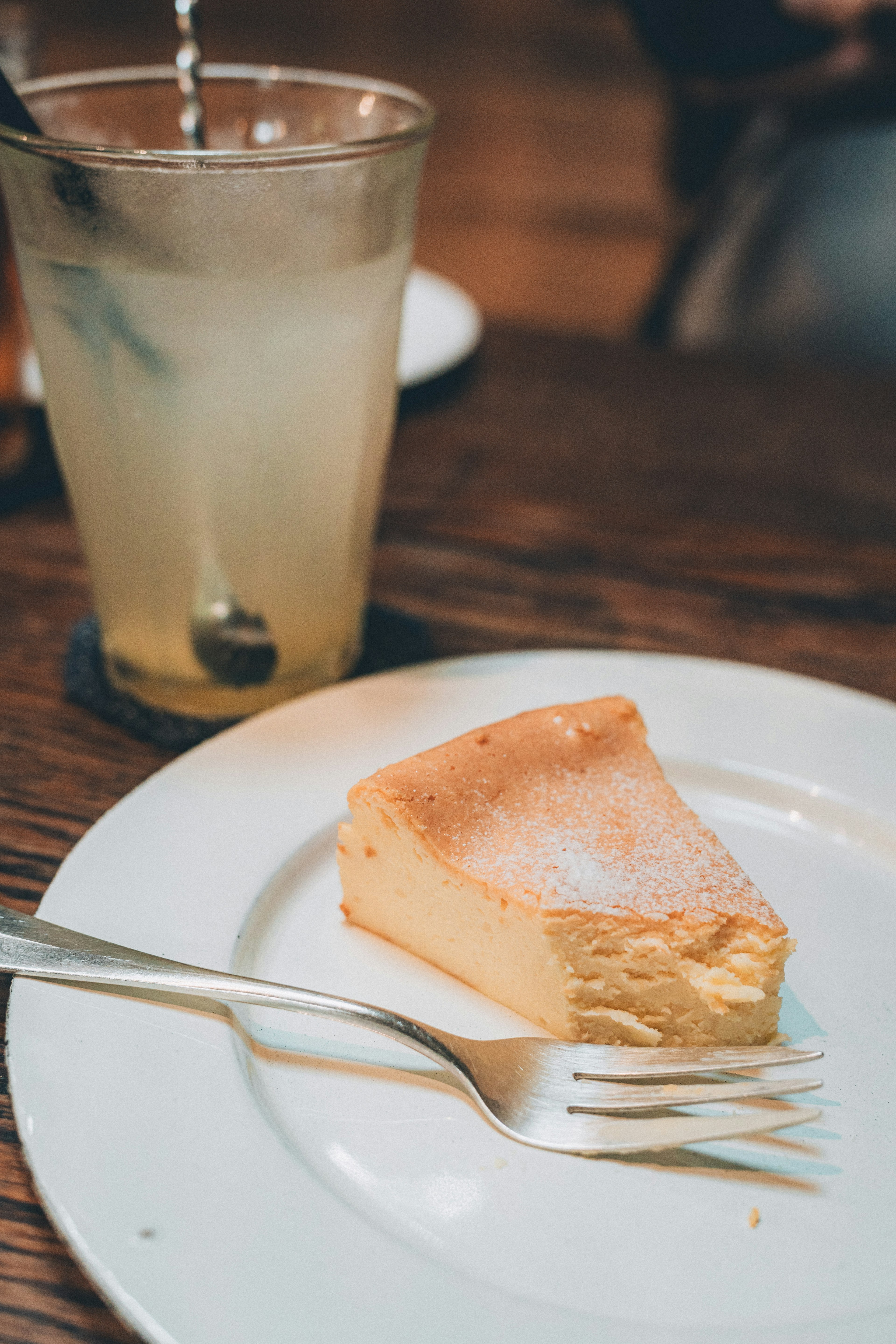 Kue keju krim di atas piring putih dengan garpu dan segelas limun