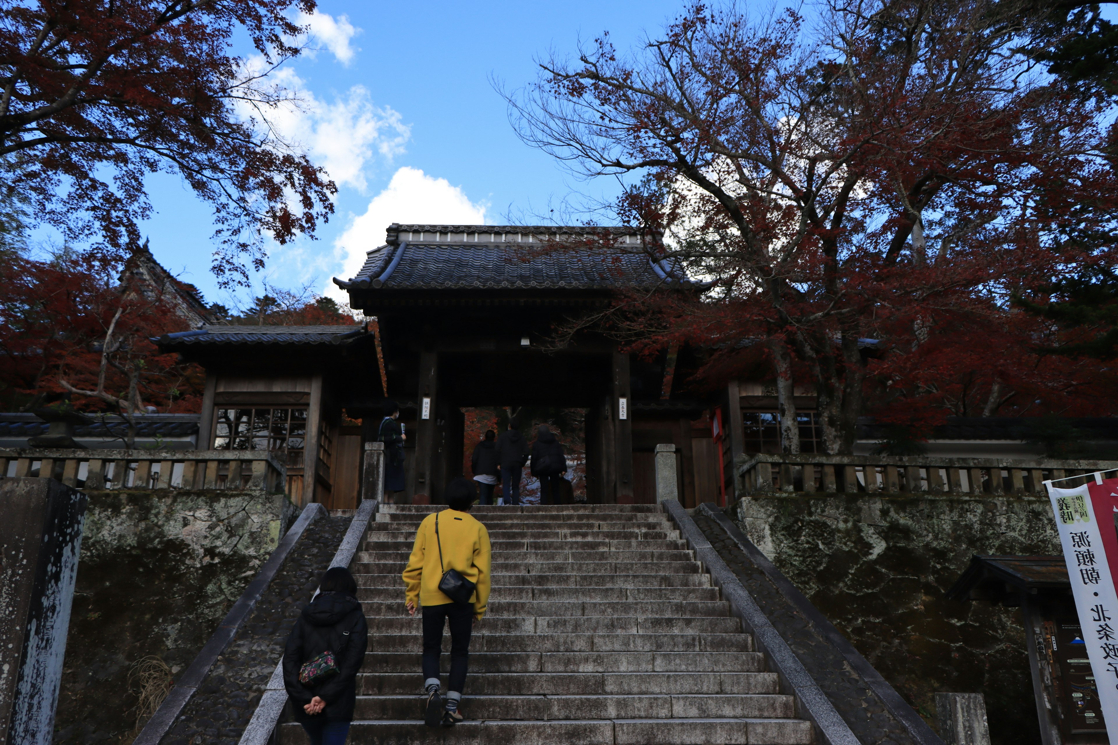 Entrance of a shrine with people climbing stairs surrounded by autumn leaves and blue sky