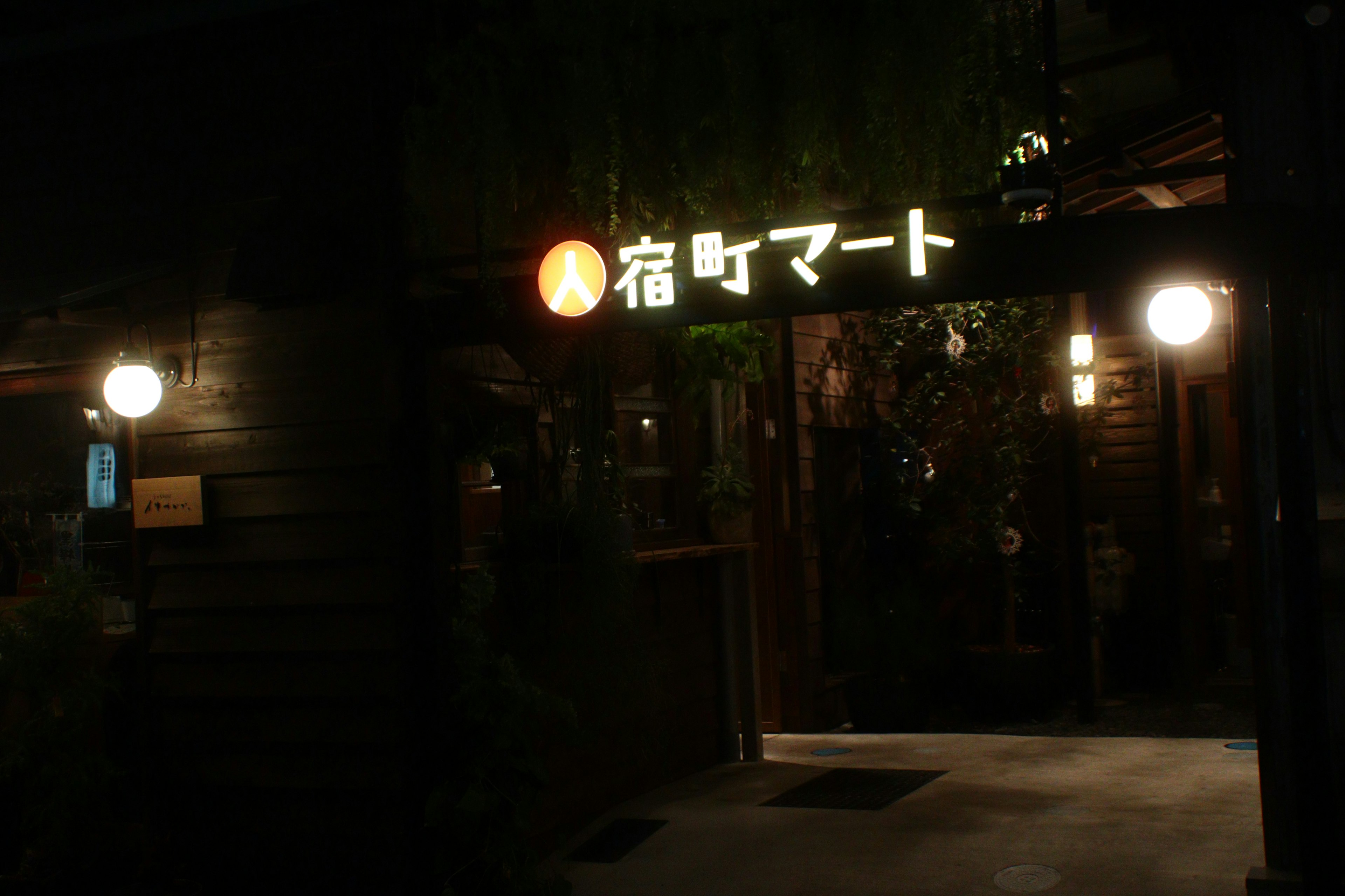 Store entrance at night featuring a bright sign that reads '宿泊マート'