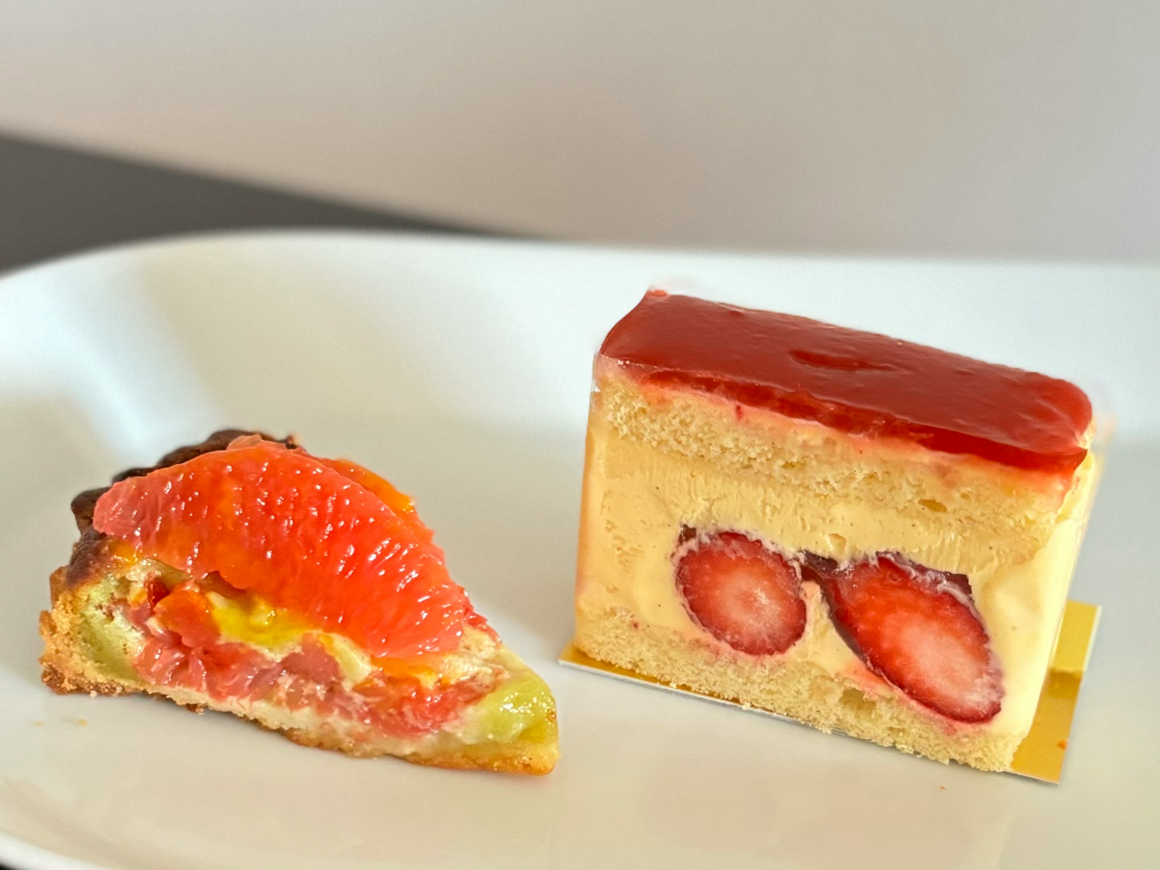 An image of two desserts on a white plate. On the left is a fruit tart topped with citrus. On the right is a cream cake featuring strawberries