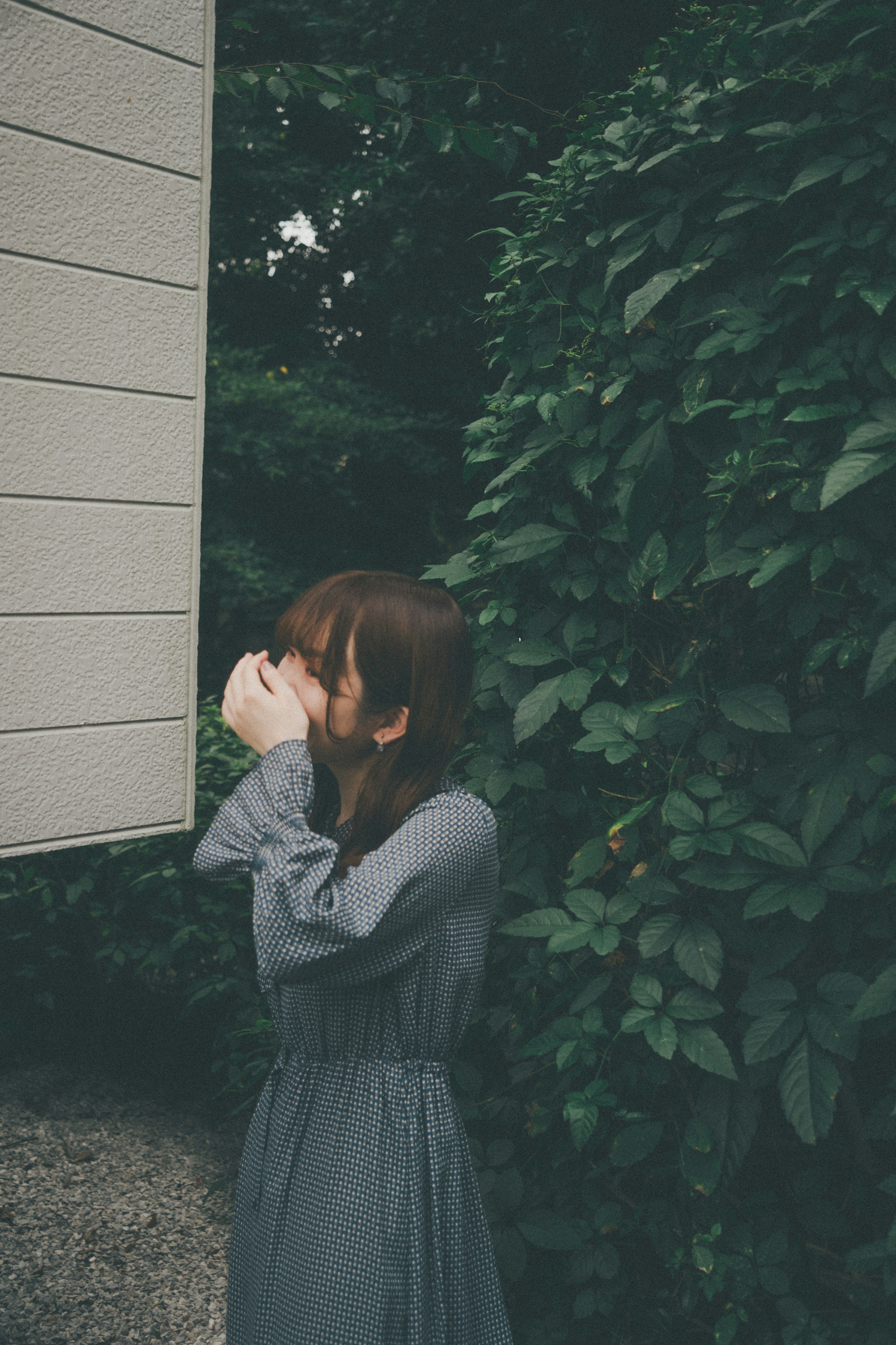 A woman in a gray dress holding a camera and hiding her face surrounded by green foliage