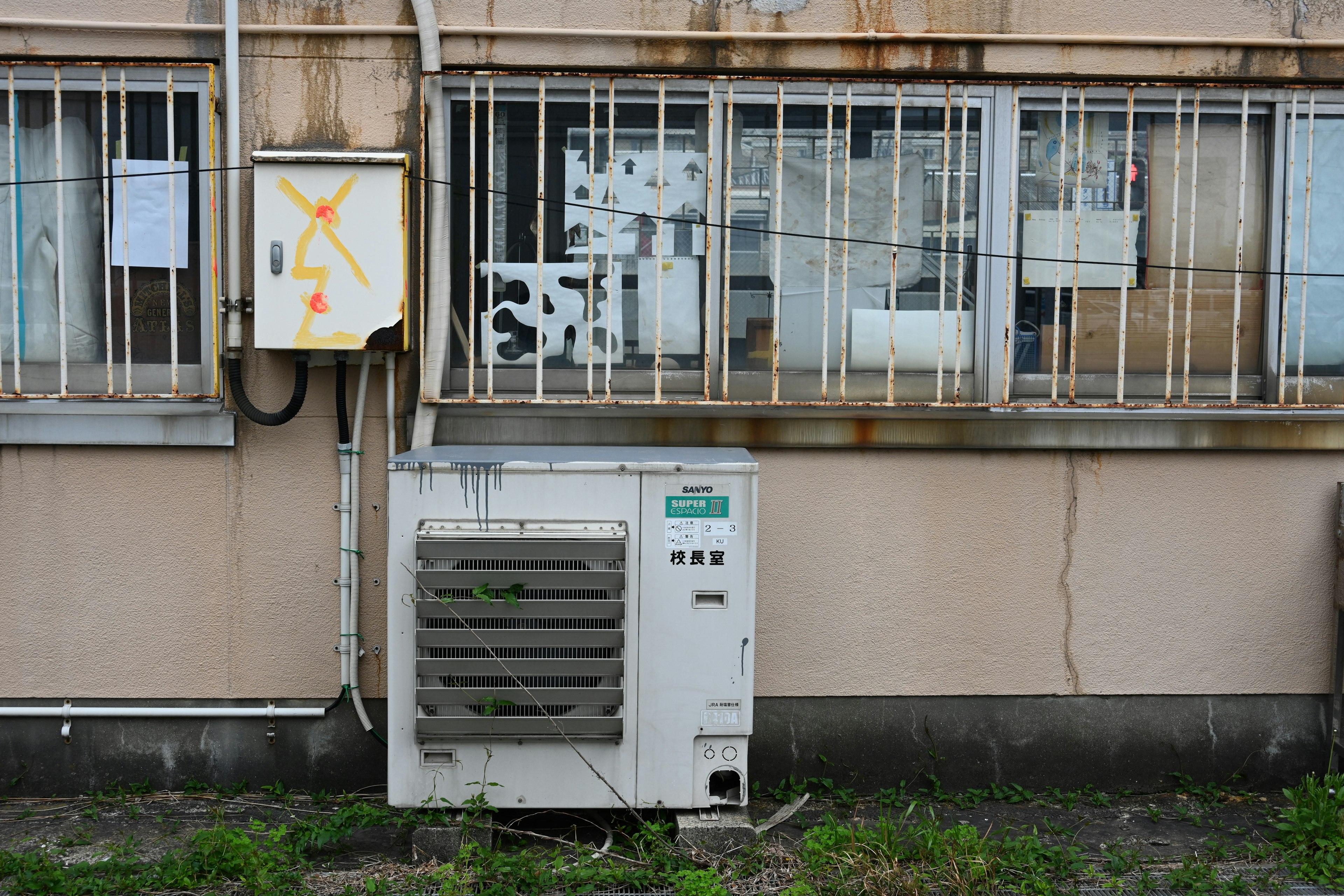 Air conditioning unit and electrical box on the exterior of an old building Rusty metal fence and overgrown grass