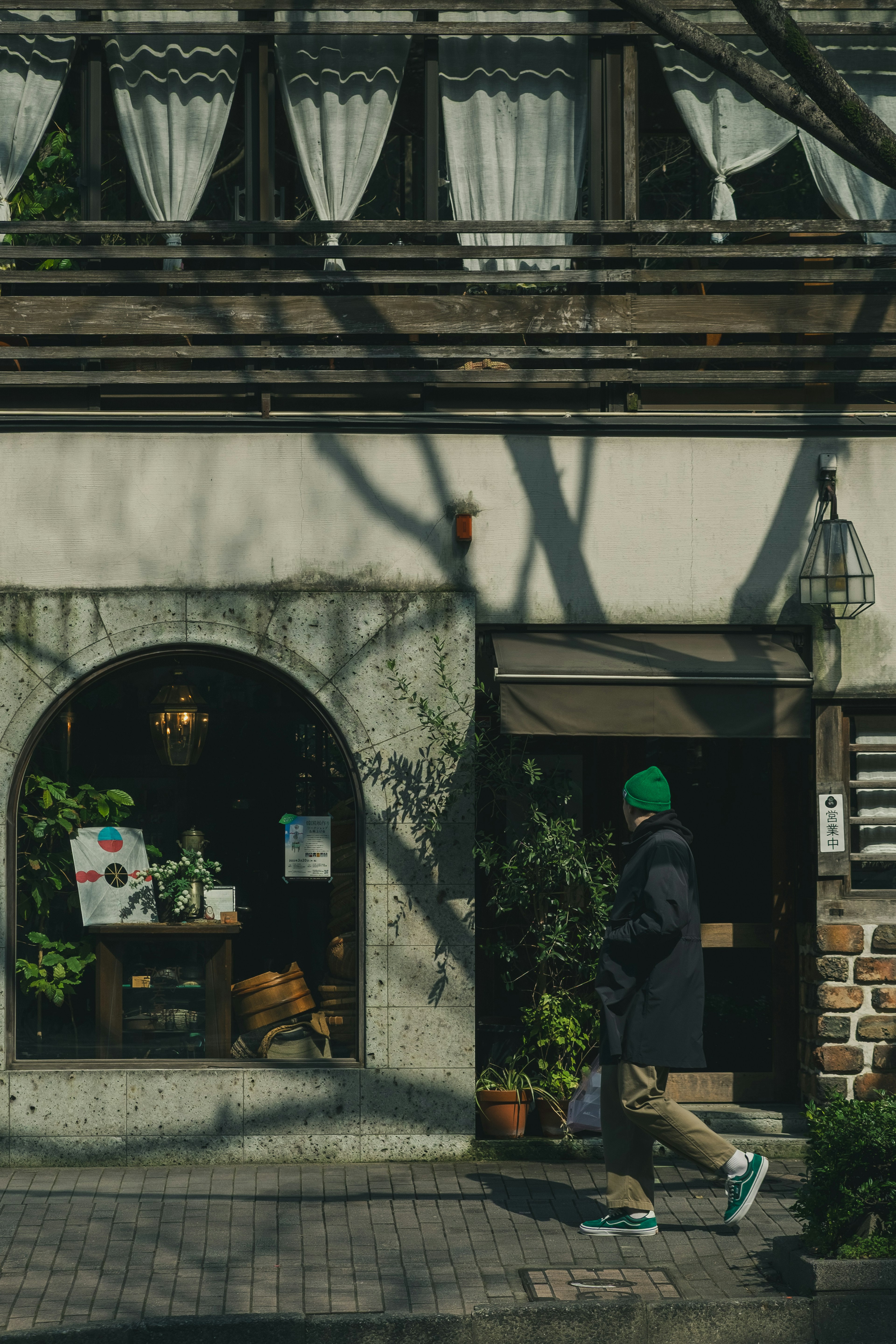 緑の帽子をかぶった人物が店舗の前を通り過ぎる風景 店舗には観葉植物とカーテンが見える