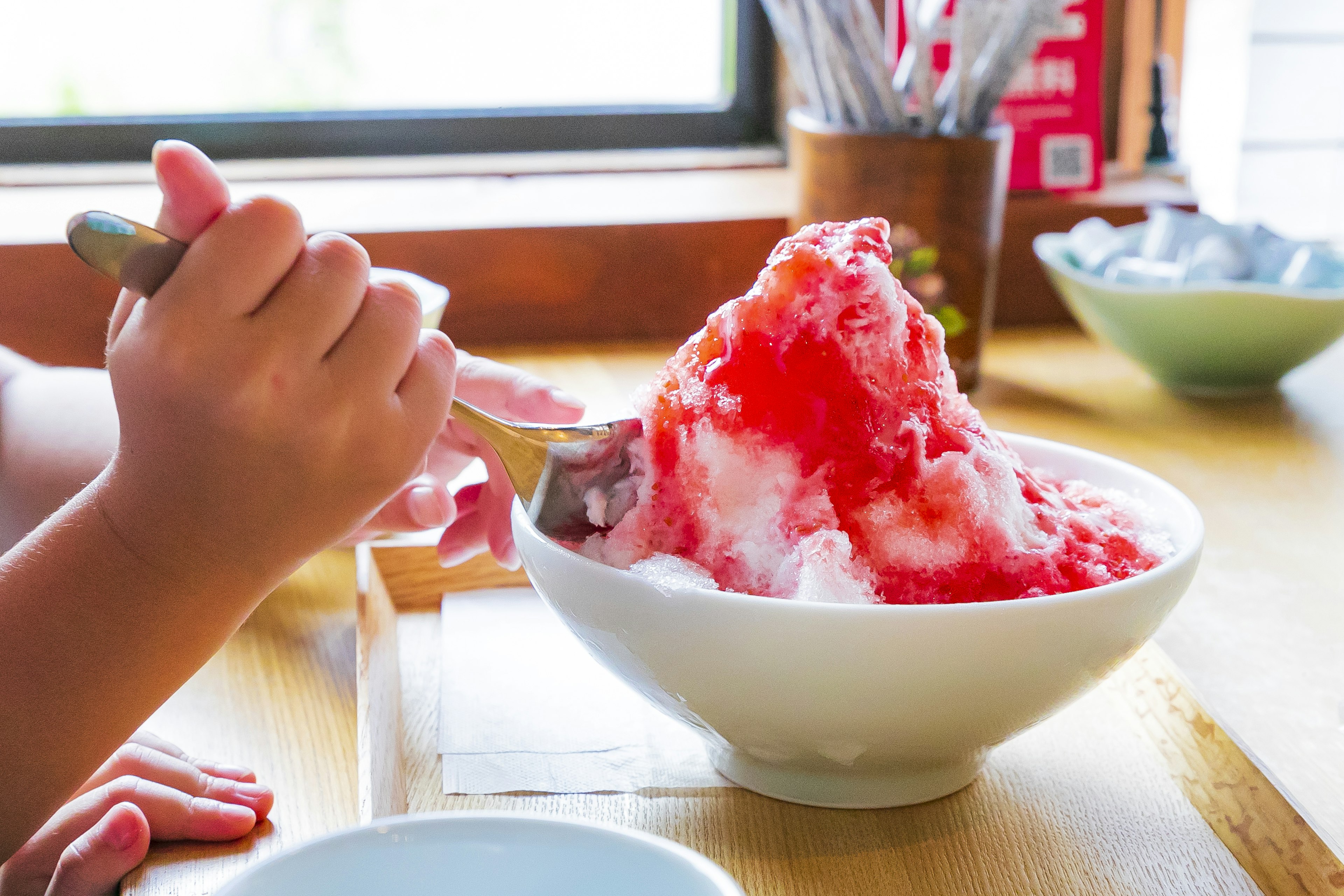 Hand hält einen Löffel mit einer Schüssel geriebenem Eis, bedeckt mit rotem Sirup