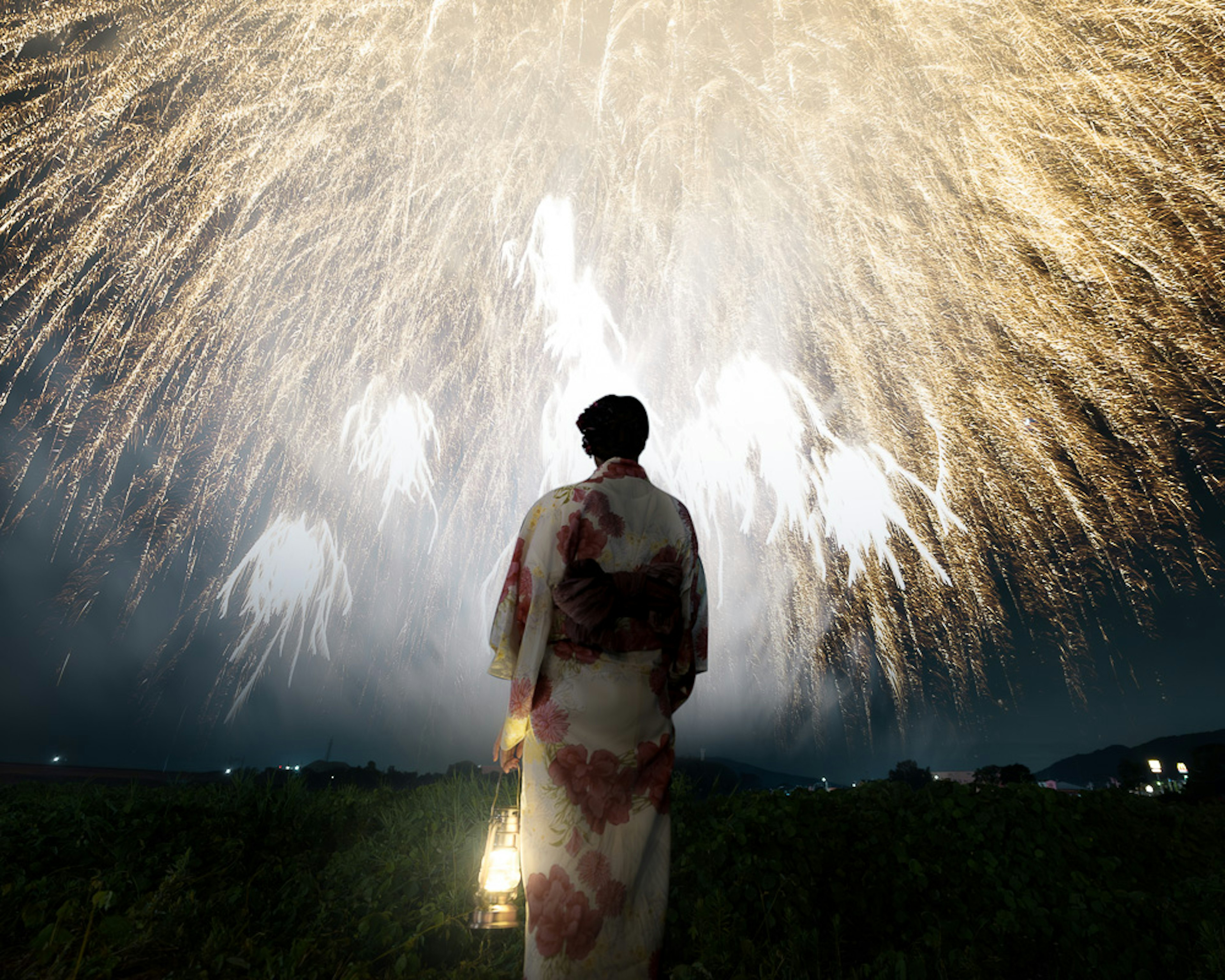 A woman in a kimono holding a lantern stands against a backdrop of fireworks in the night sky