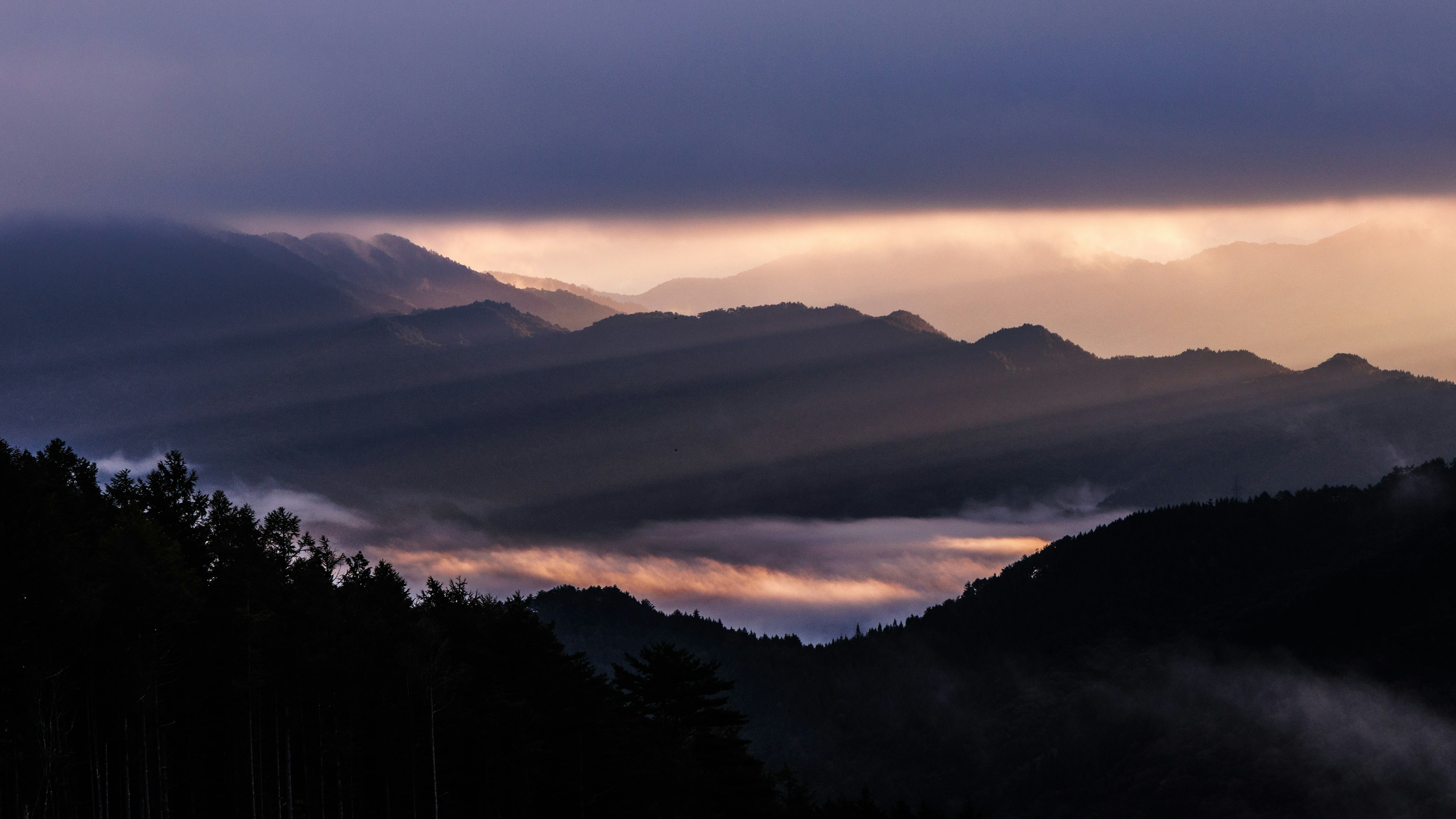 Pemandangan pegunungan yang diselimuti kabut dengan langit kelam dan nuansa matahari terbenam ungu