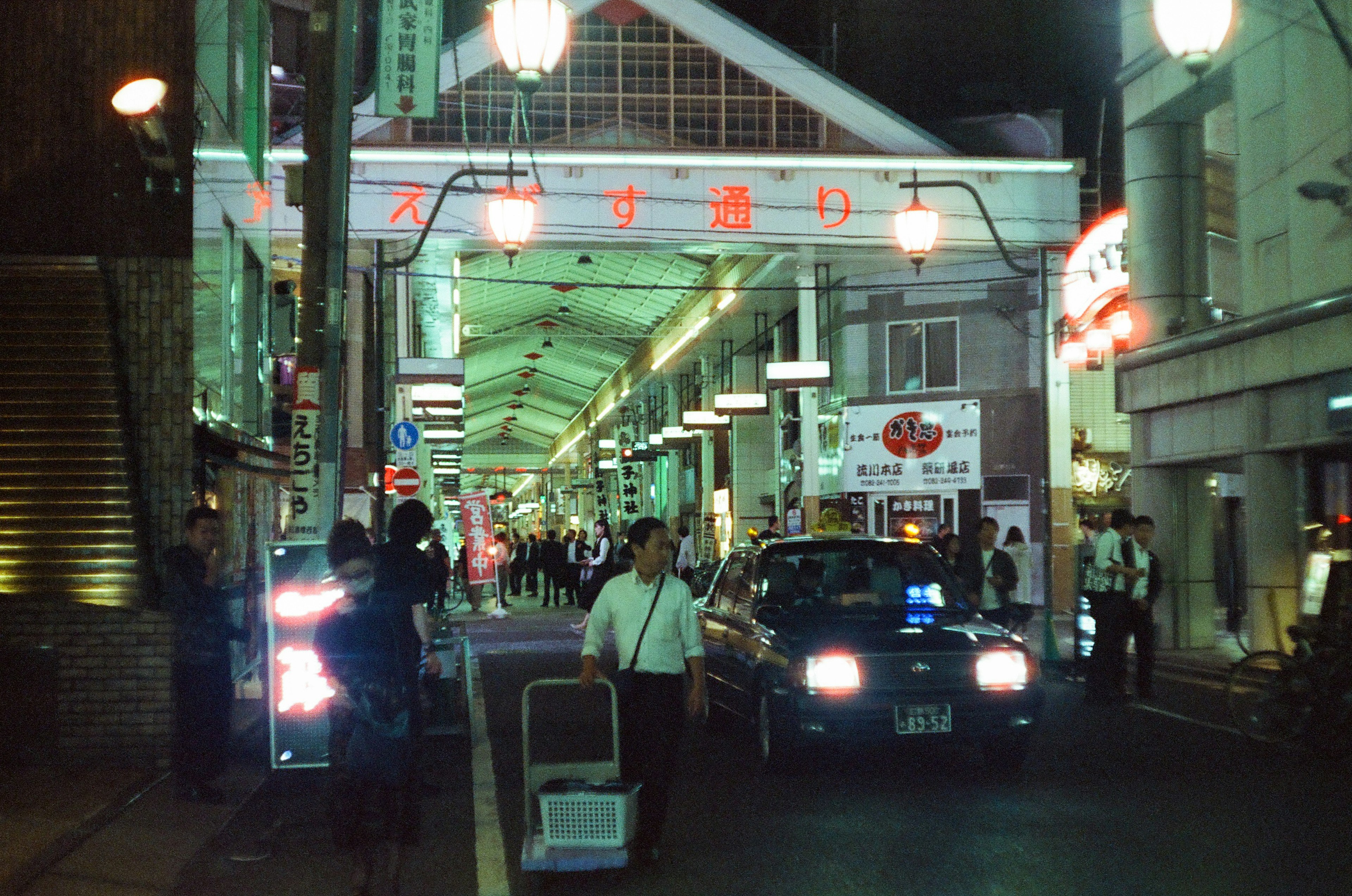 Busy nighttime street with pedestrians and a parked taxi illuminated by green streetlights