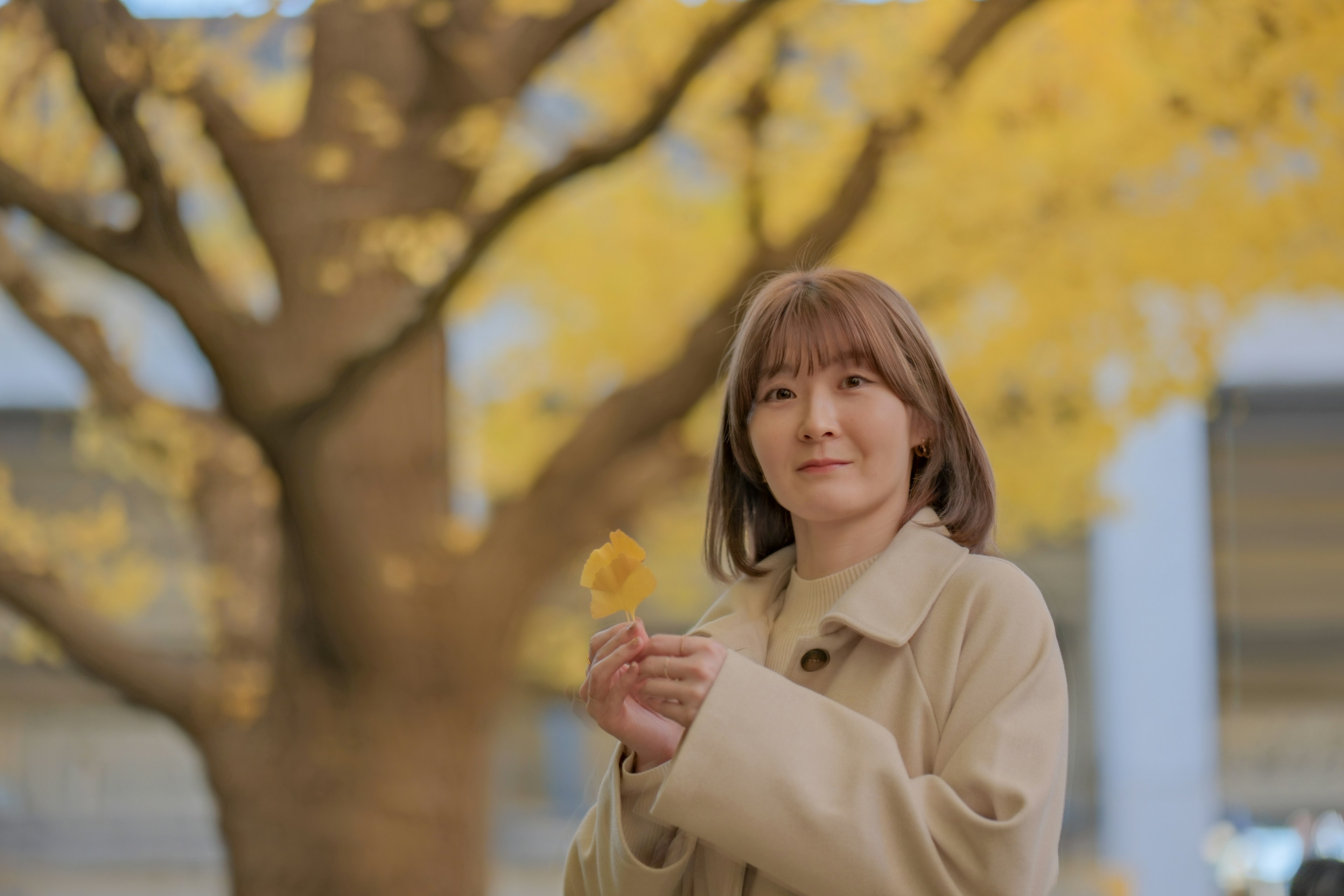 女性が黄色の花を持ち、背景に黄色い葉の木がある