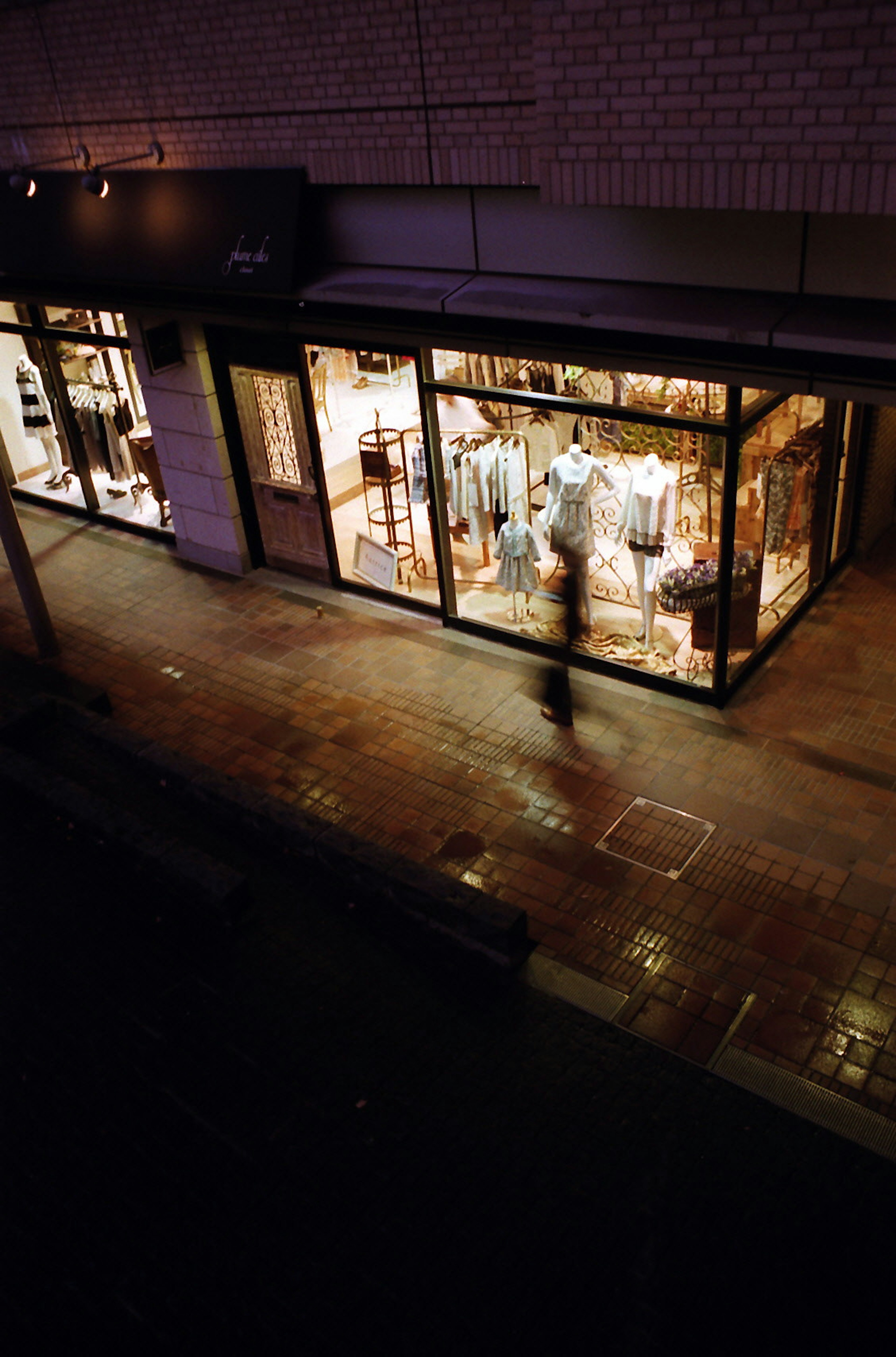 Beleuchtete Schaufenster eines Bekleidungsgeschäfts auf nassem Bürgersteig mit schattenhafter Figur