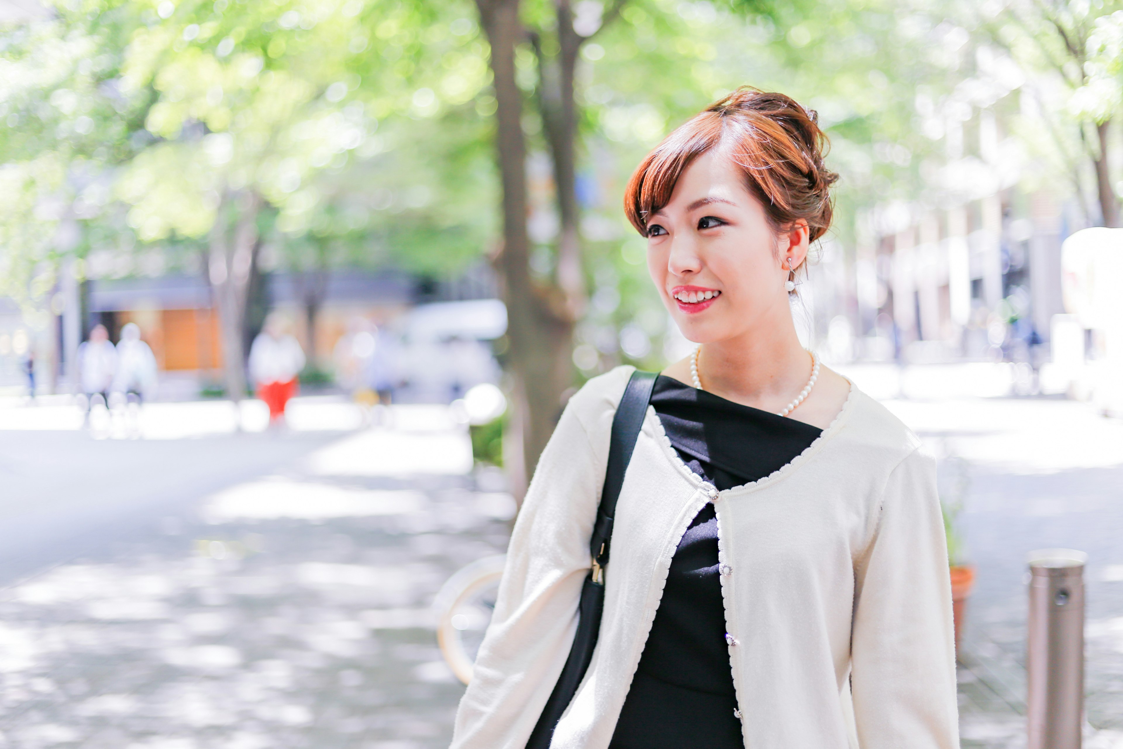 Smiling woman in a park with green trees and bright sunlight wearing casual clothing and a black handbag