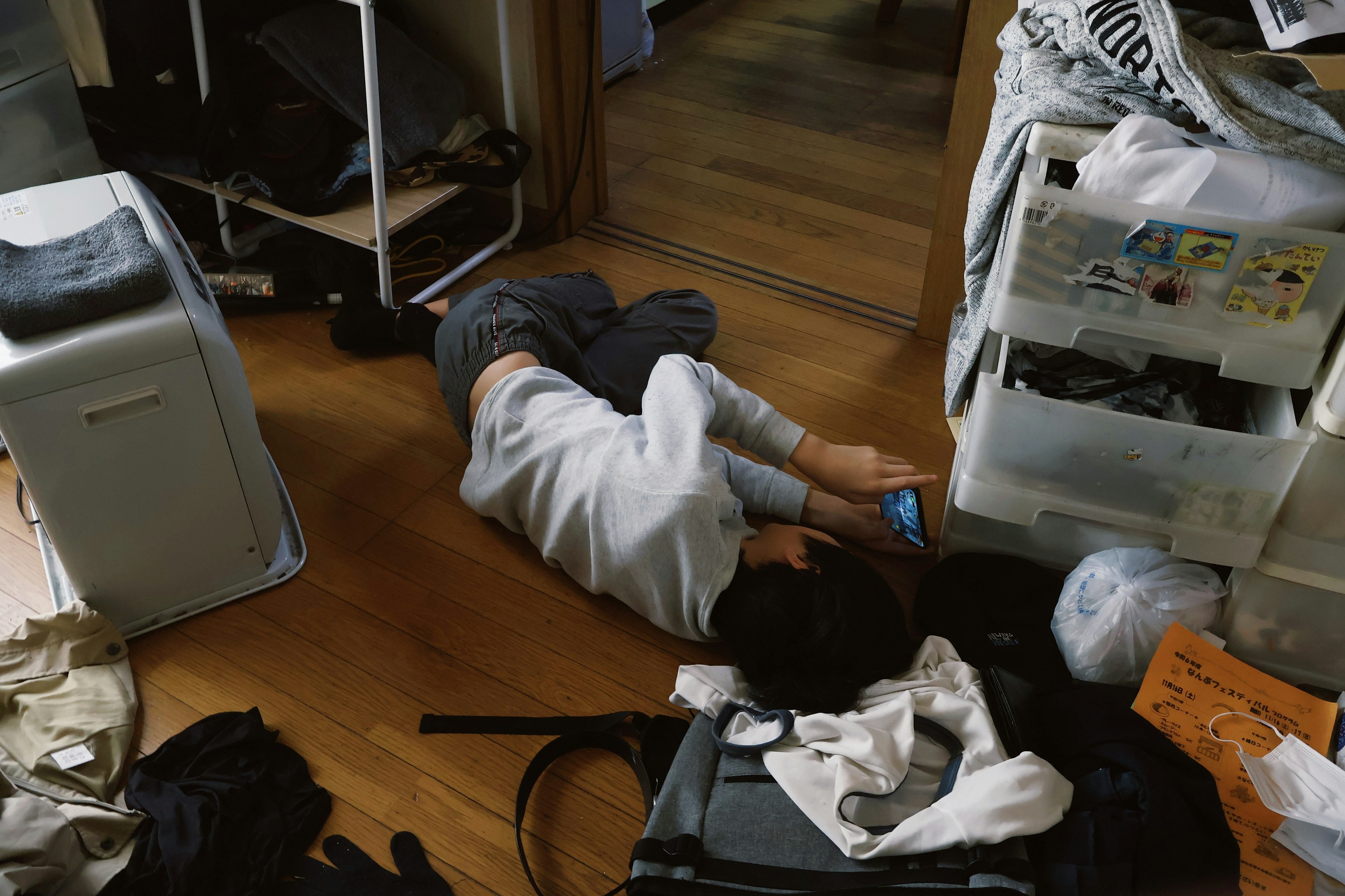 A young person lying on the floor of a messy room using a smartphone