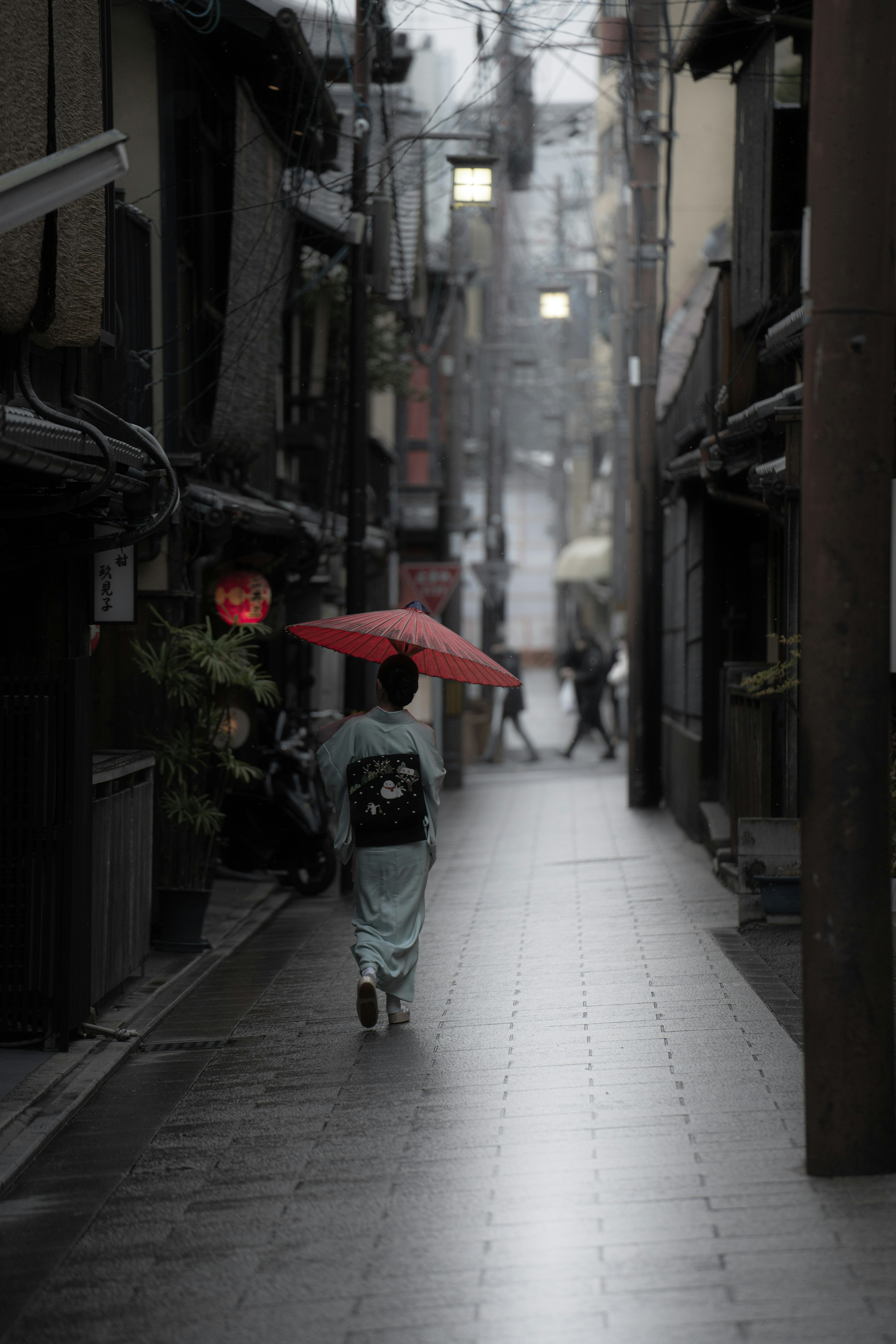 Eine Person im Kimono, die mit einem roten Regenschirm durch eine enge Gasse geht