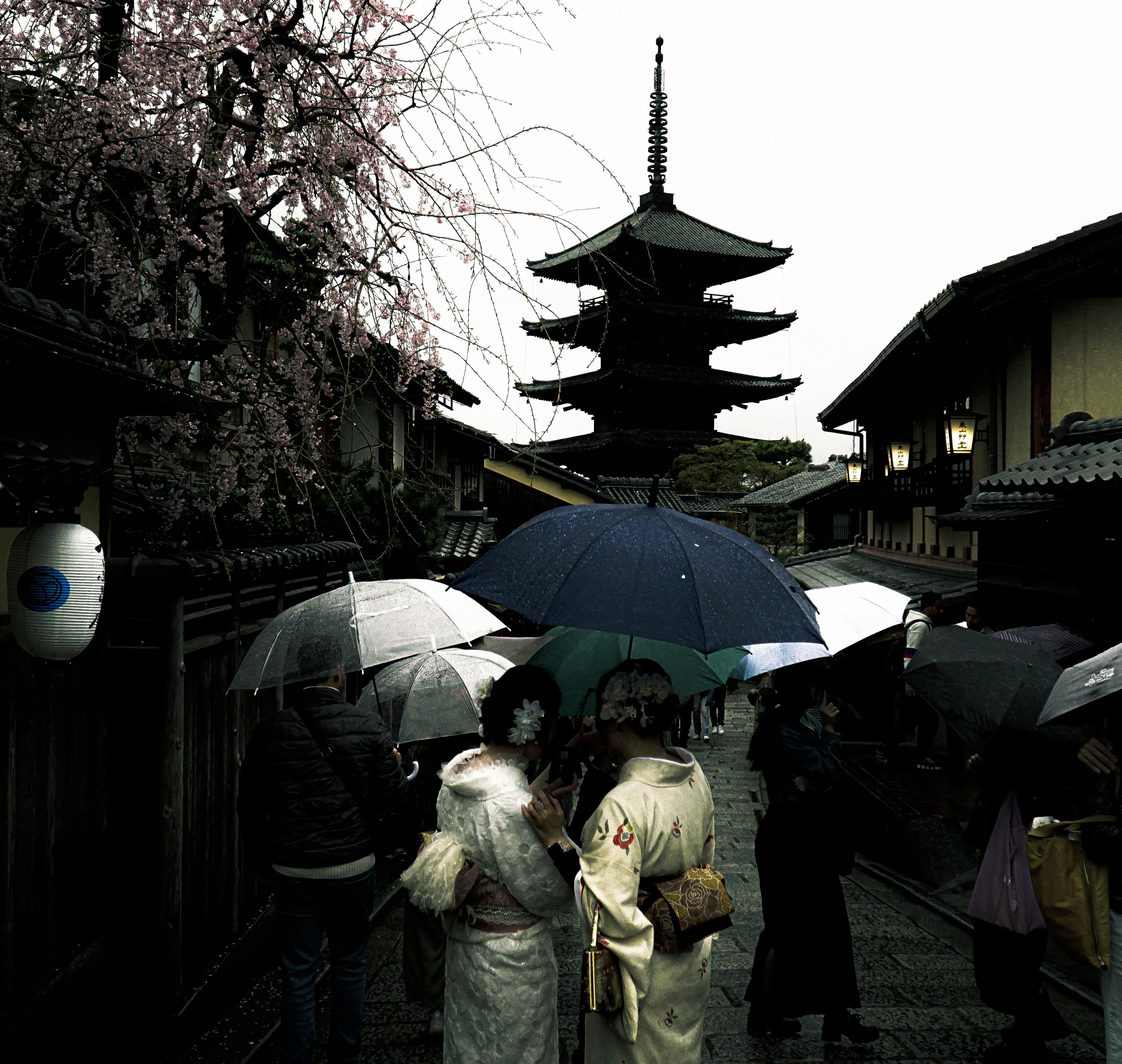 雨の中で傘を持つ着物姿の人々と五重塔が見える古い街並み