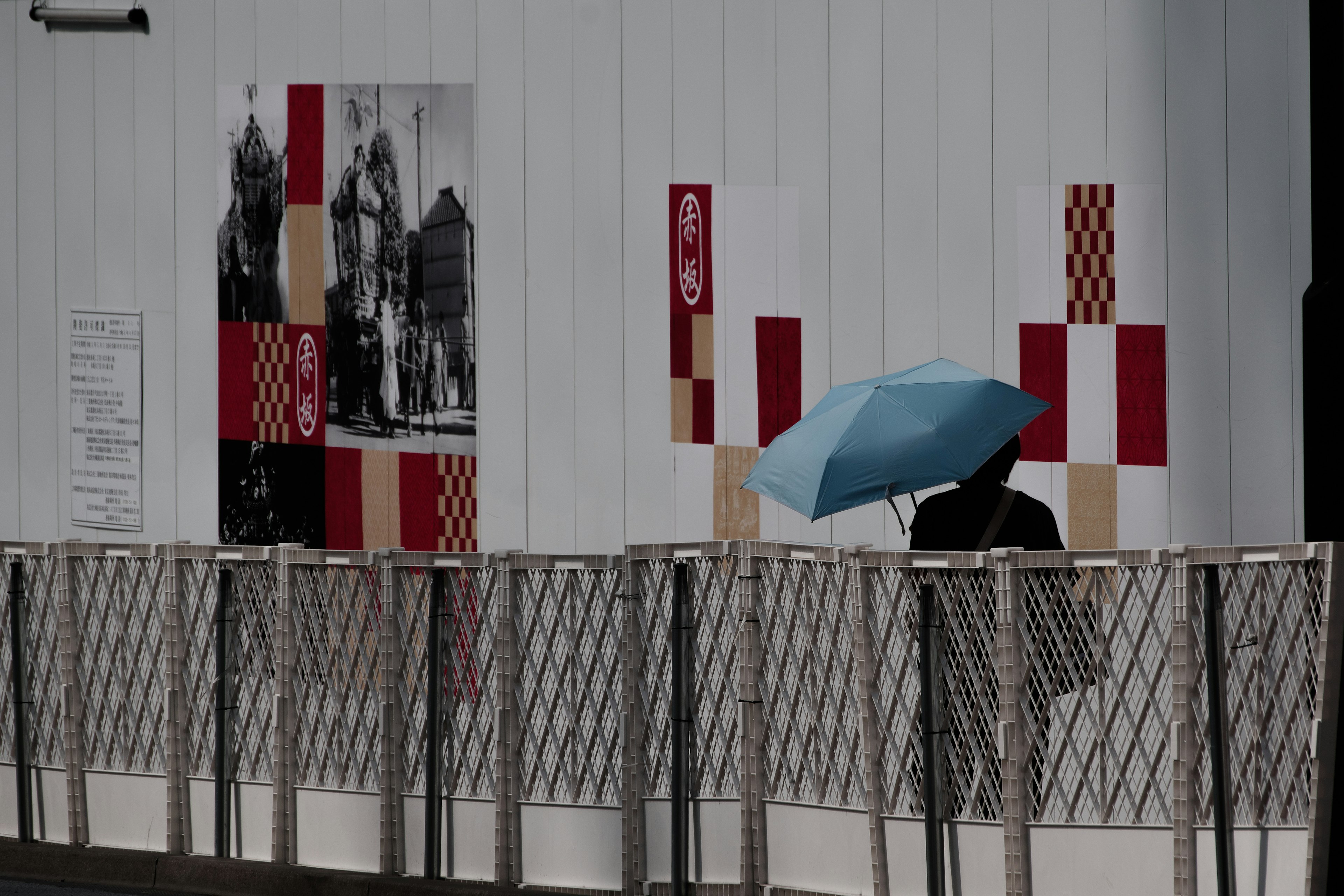 Une personne avec un parapluie bleu marchant devant un mur blanc avec des décorations rouges et blanches et une photo