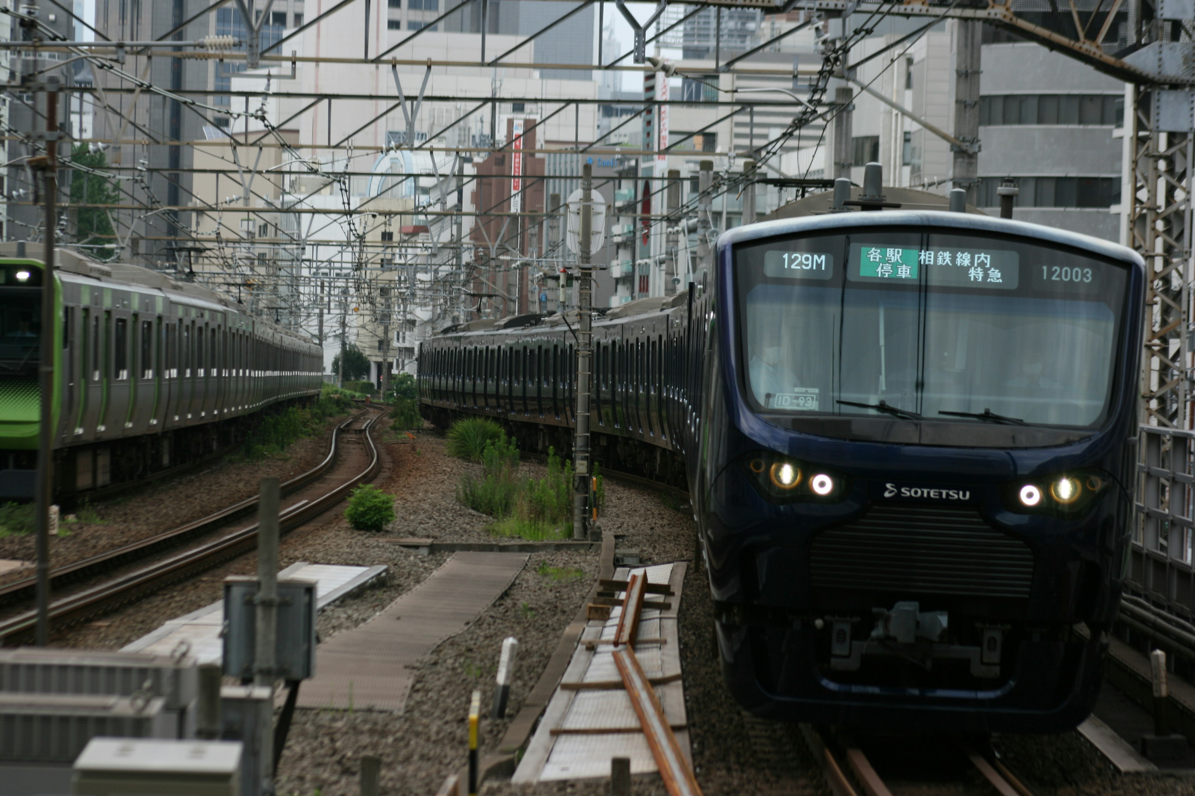 Escena ferroviaria urbana con un tren negro y un tren verde en vías paralelas con rascacielos de fondo