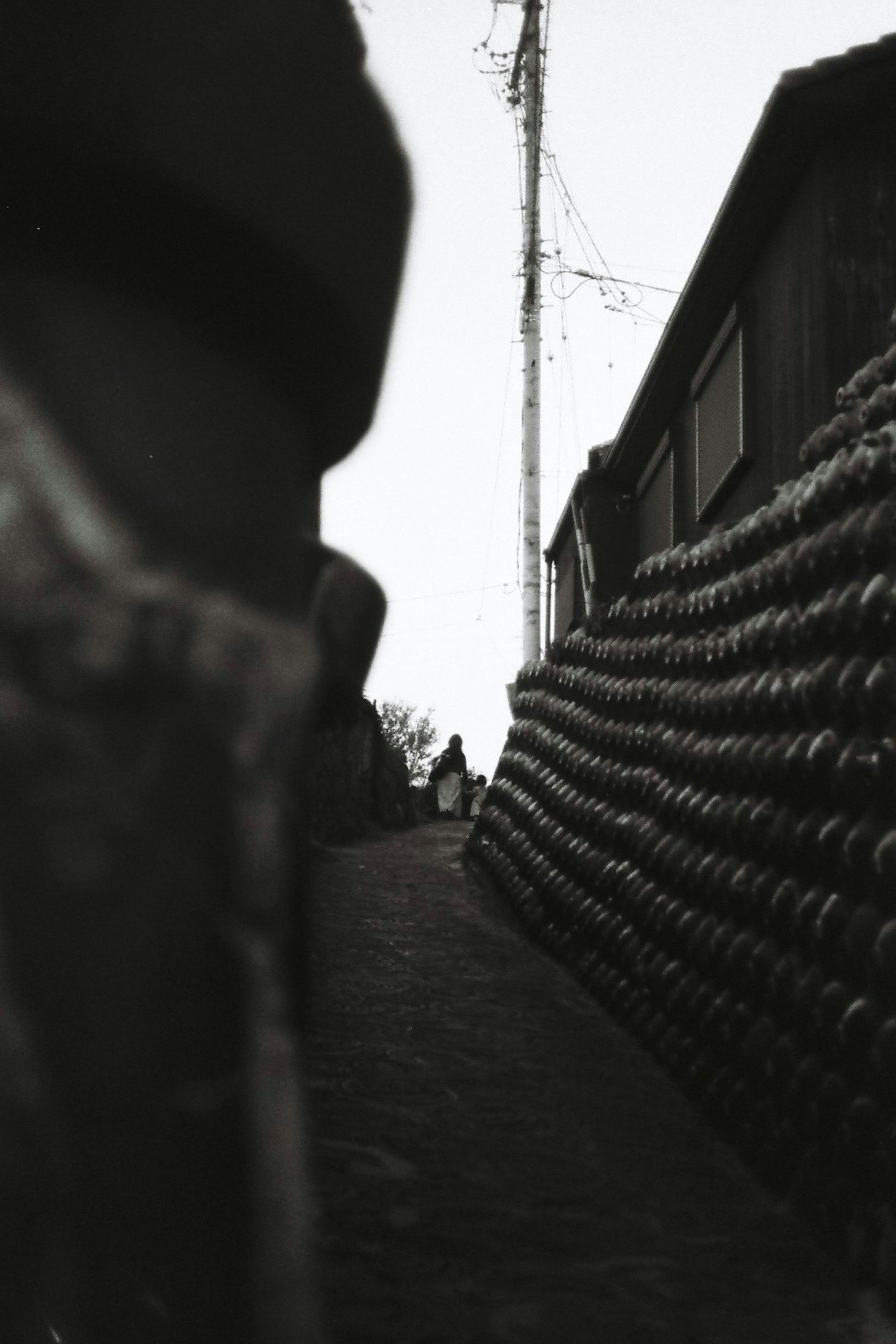 Black and white photo of a narrow alley featuring cylindrical objects stacked on a wall and a distant figure