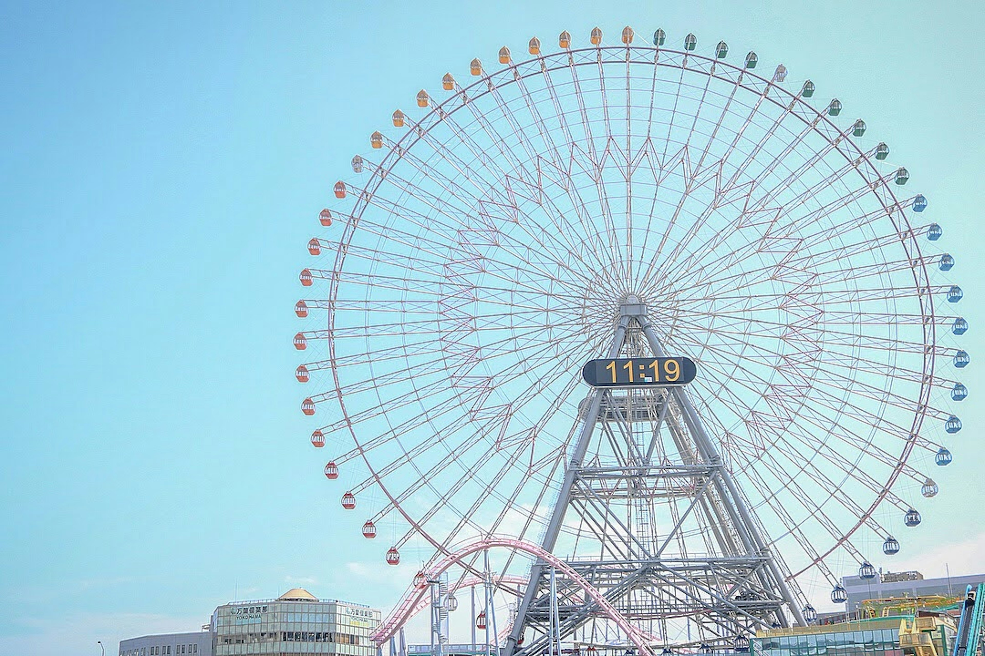 Una grande ruota panoramica contro un cielo blu chiaro con elementi di parco divertimenti visibili