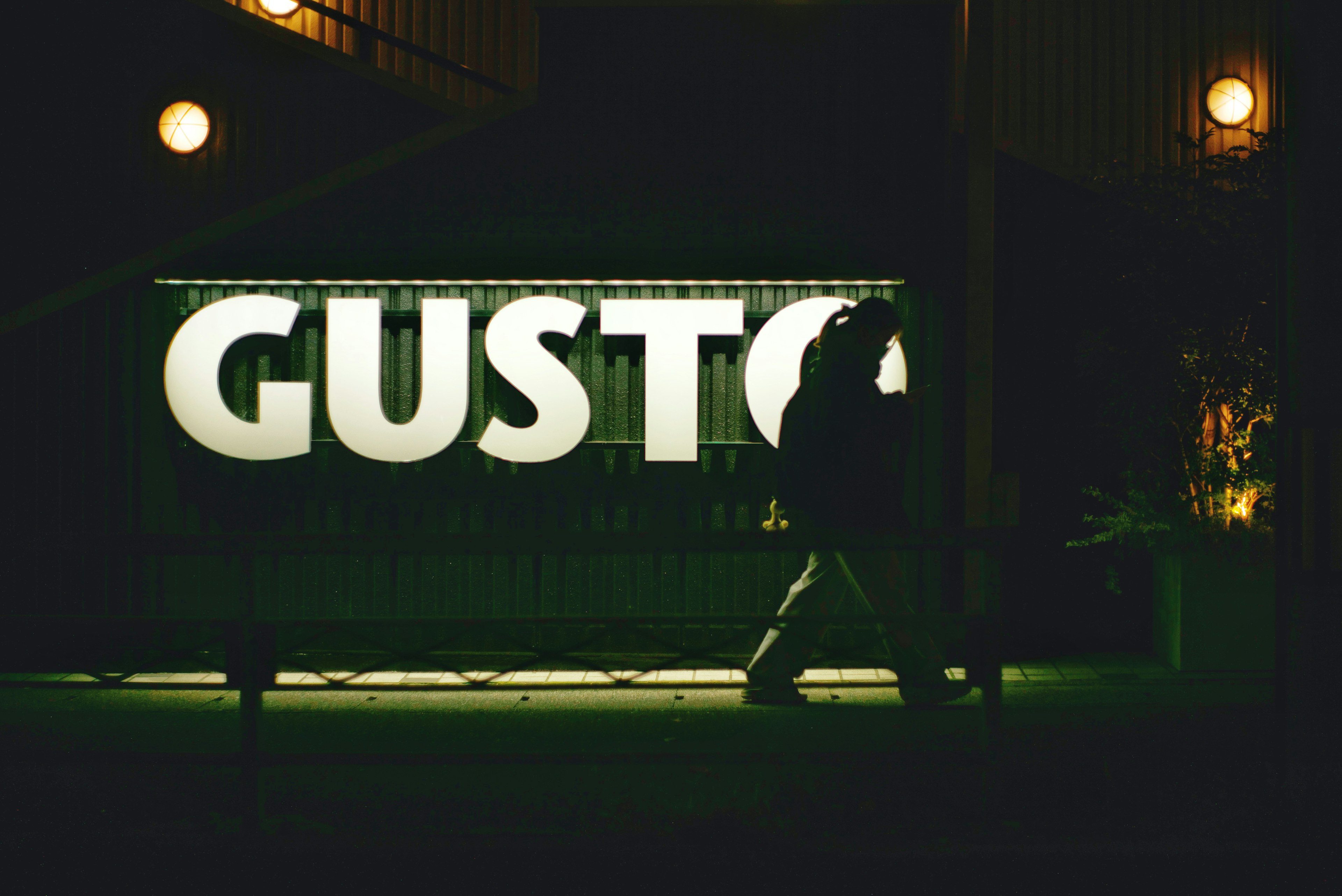 A silhouette walking past a GUSTO sign at night