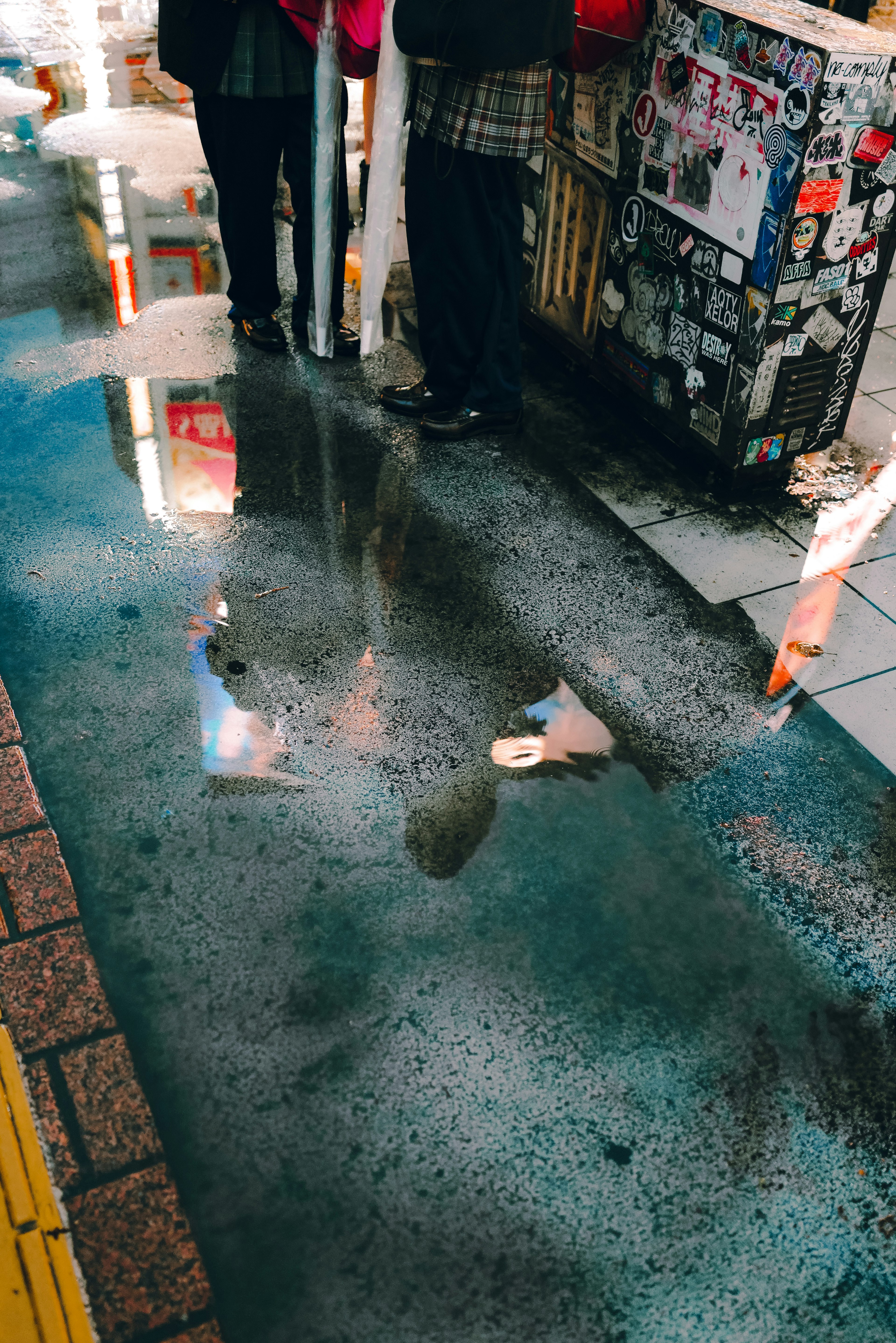 Escena de esquina de calle con un charco que refleja a personas cercanas