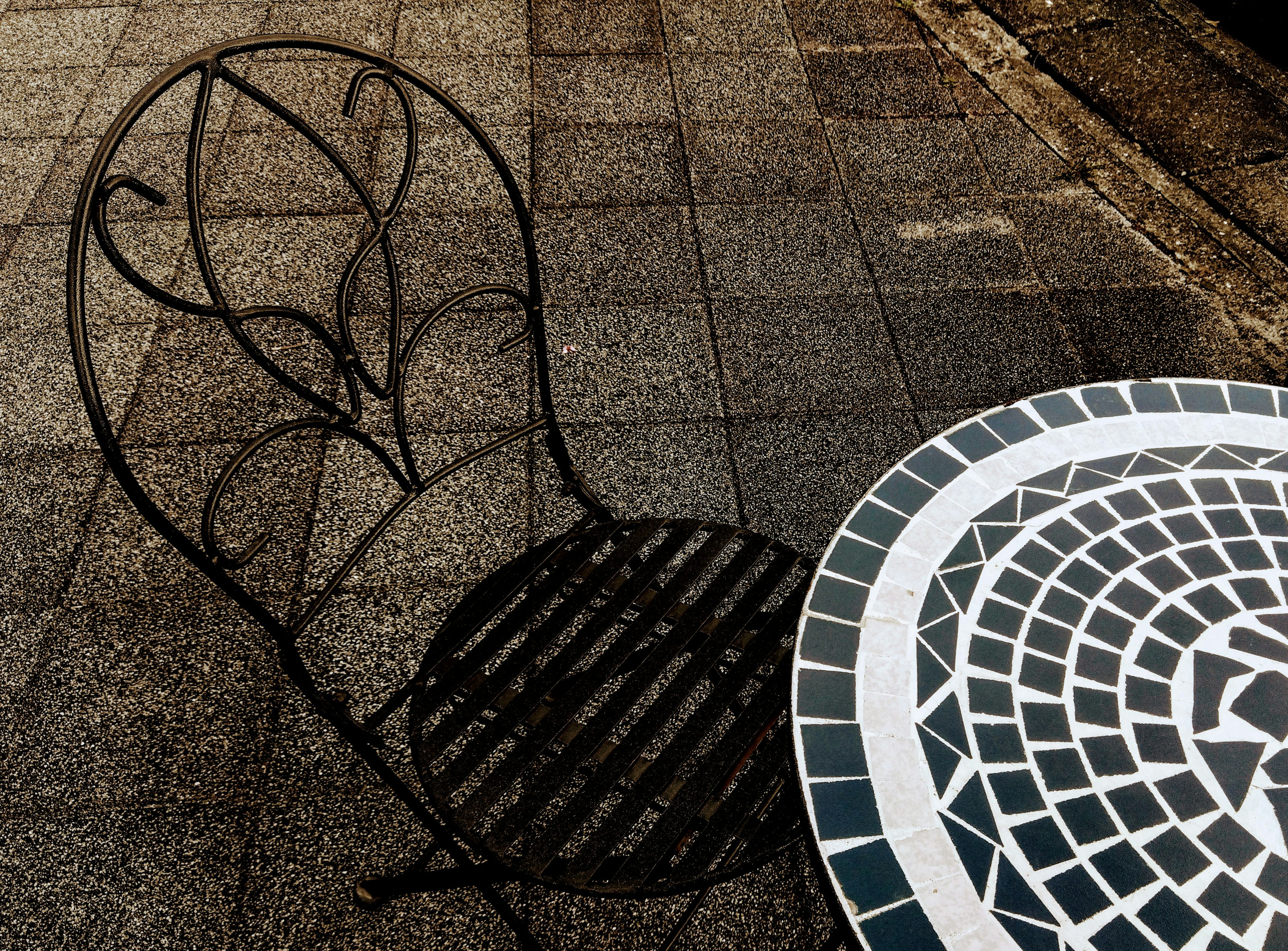 Black wrought iron chair casting a shadow beside a round mosaic table on a paved surface