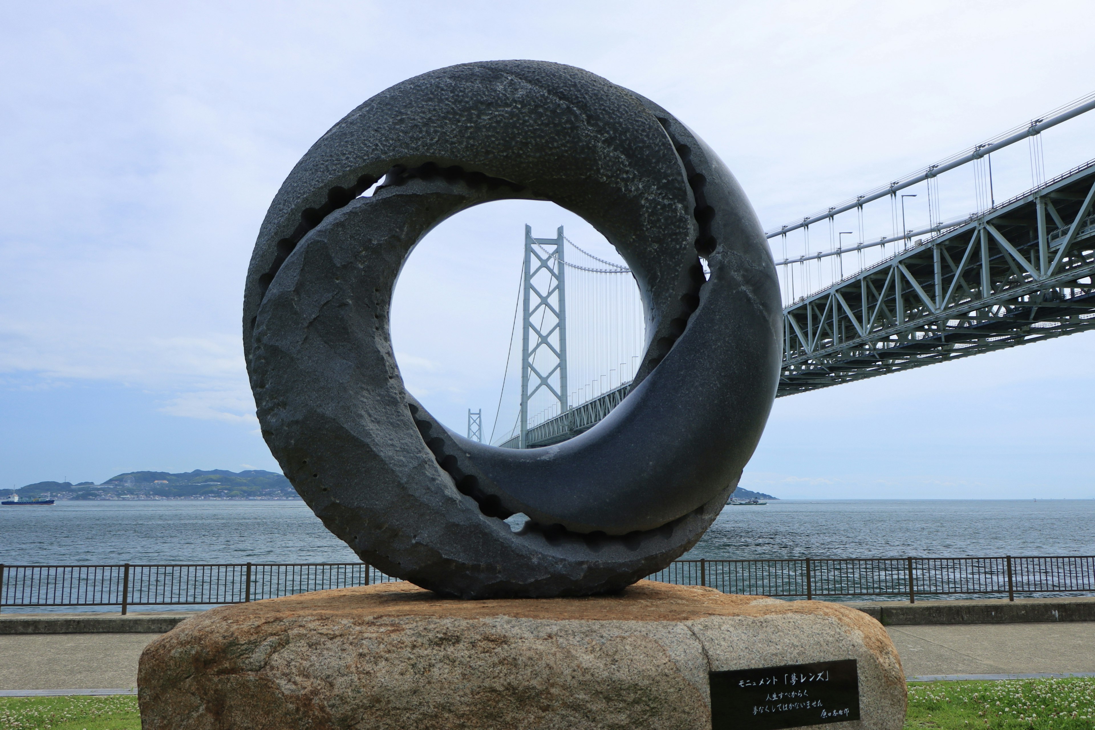 Escultura circular de piedra negra con diseño de olas cerca de un puente y el mar