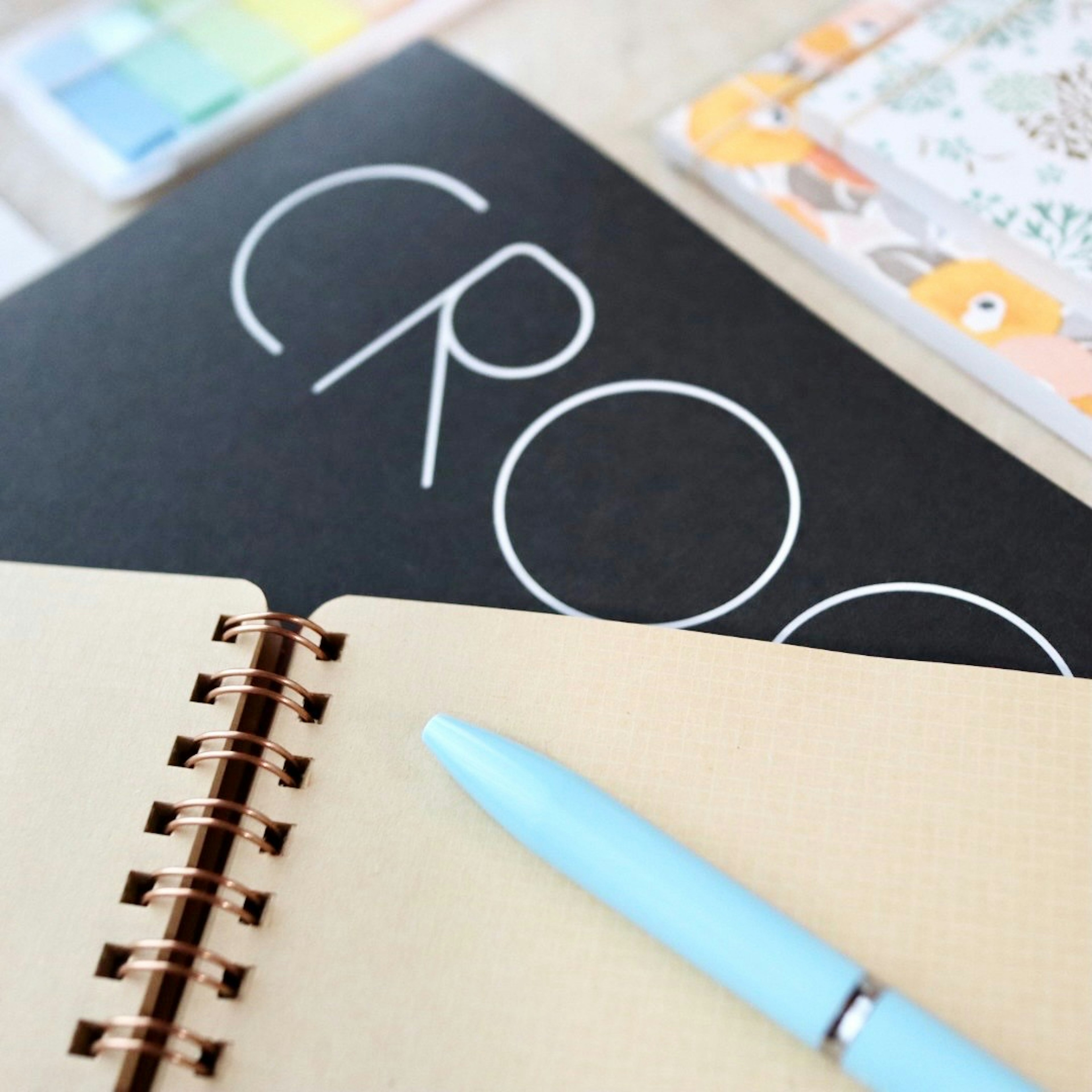 A black notebook with the word CROOK in white lettering and a light blue pen on a beige surface