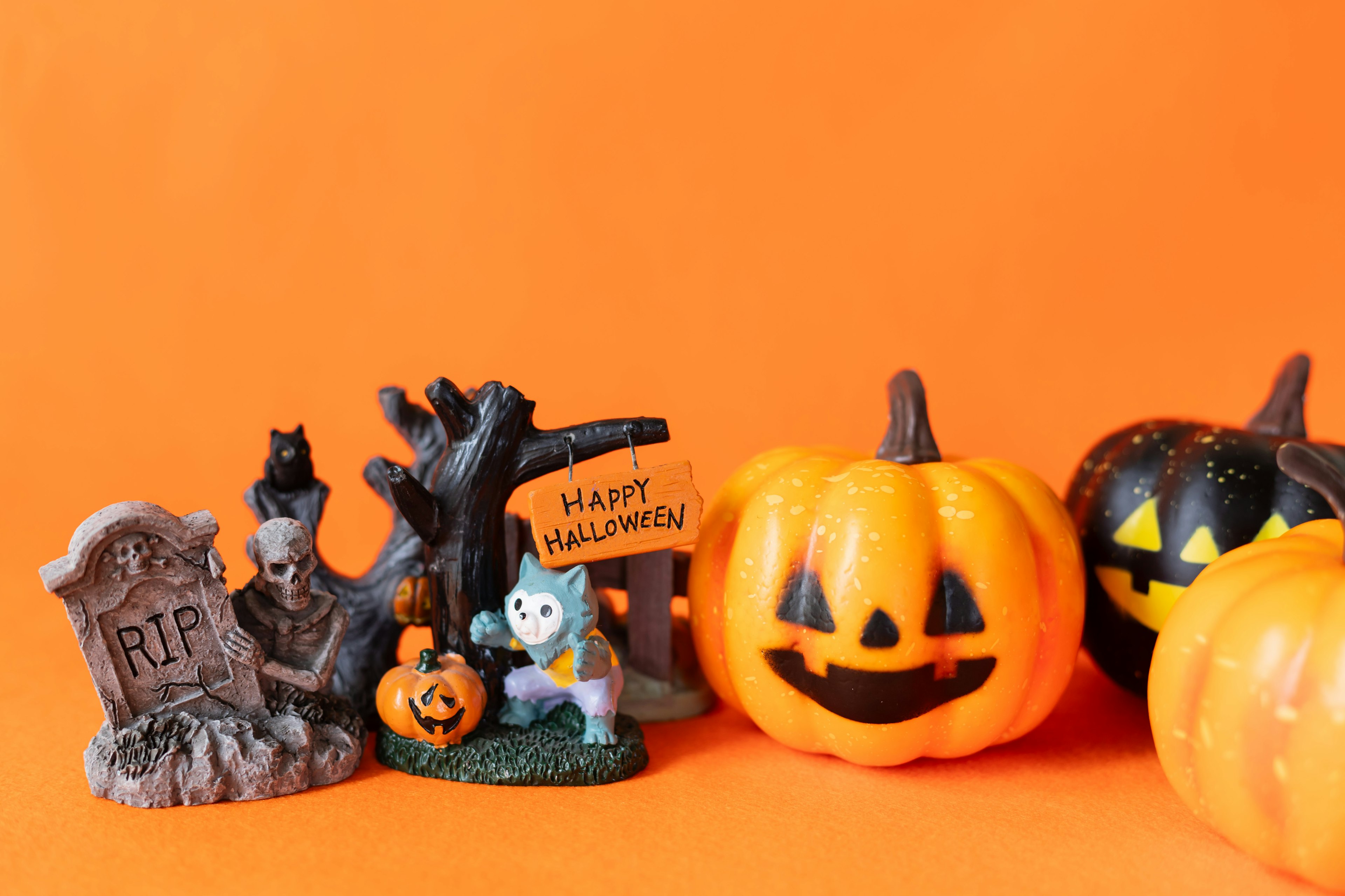 Imagen con decoraciones de Halloween sobre un fondo naranja que incluye calabazas lápidas un gato negro y un cartel de Halloween