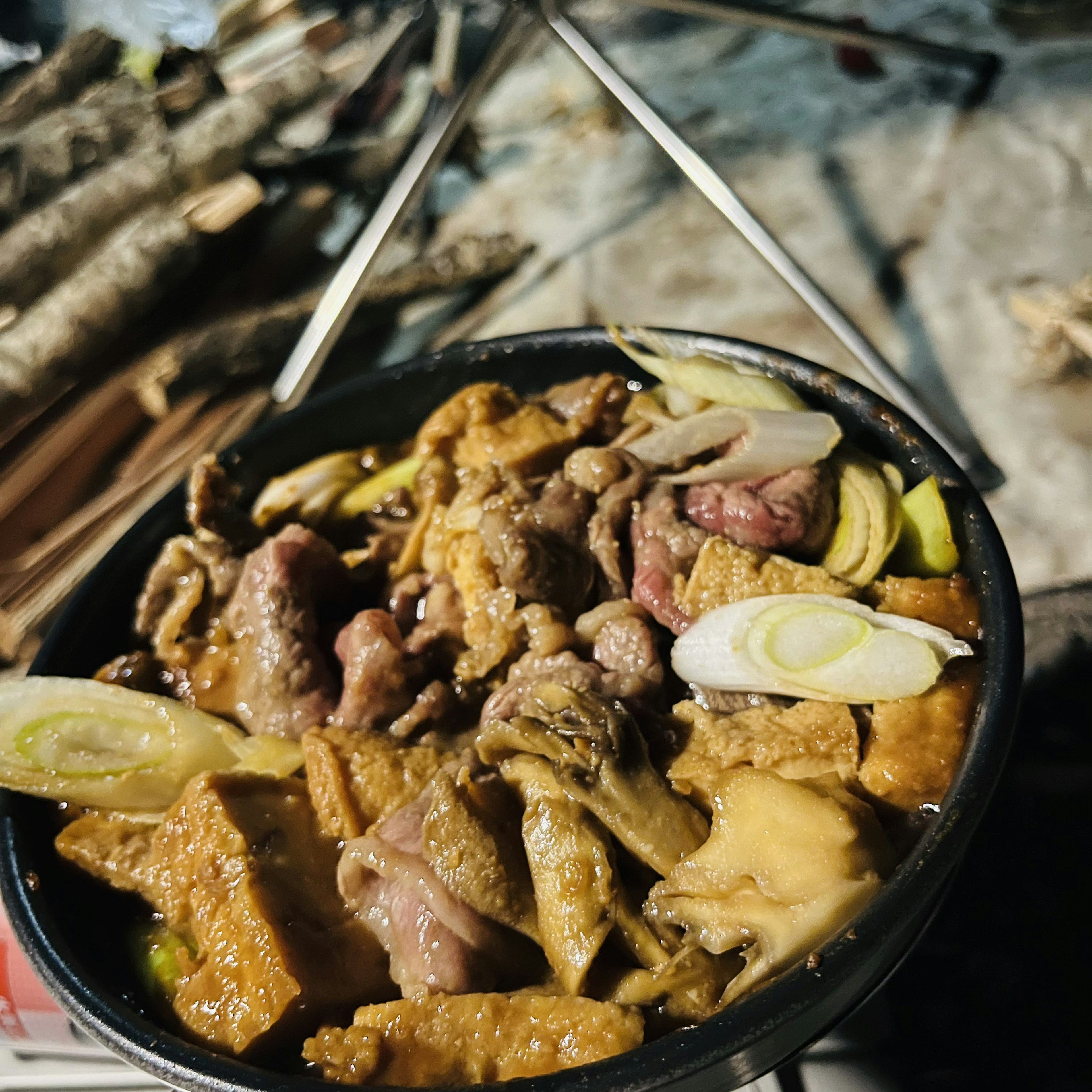 鍋の中に入った肉とネギの料理　周囲には焚き木が見える