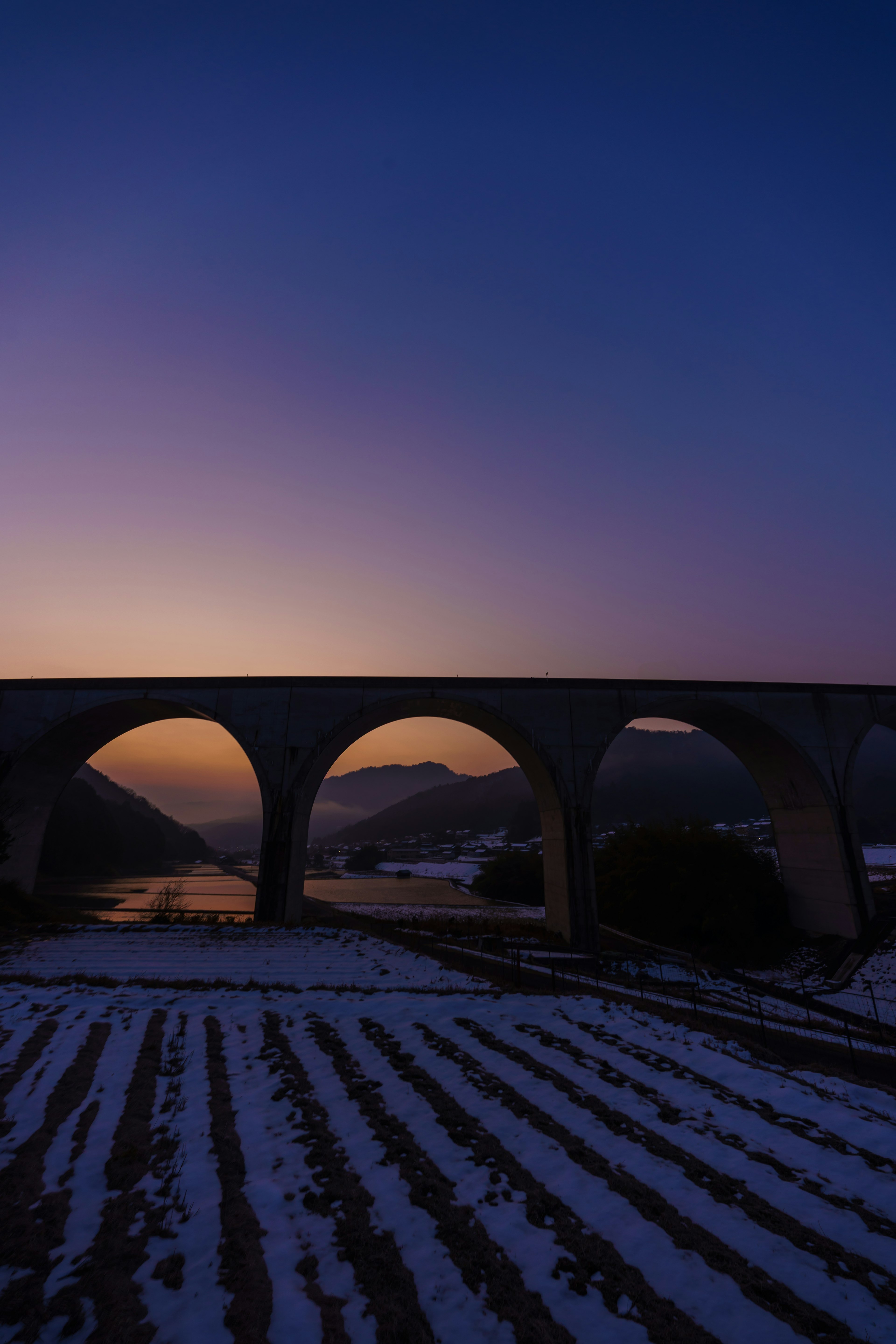 雪に覆われた畑の上に架かるアーチ型の橋と夕暮れの空