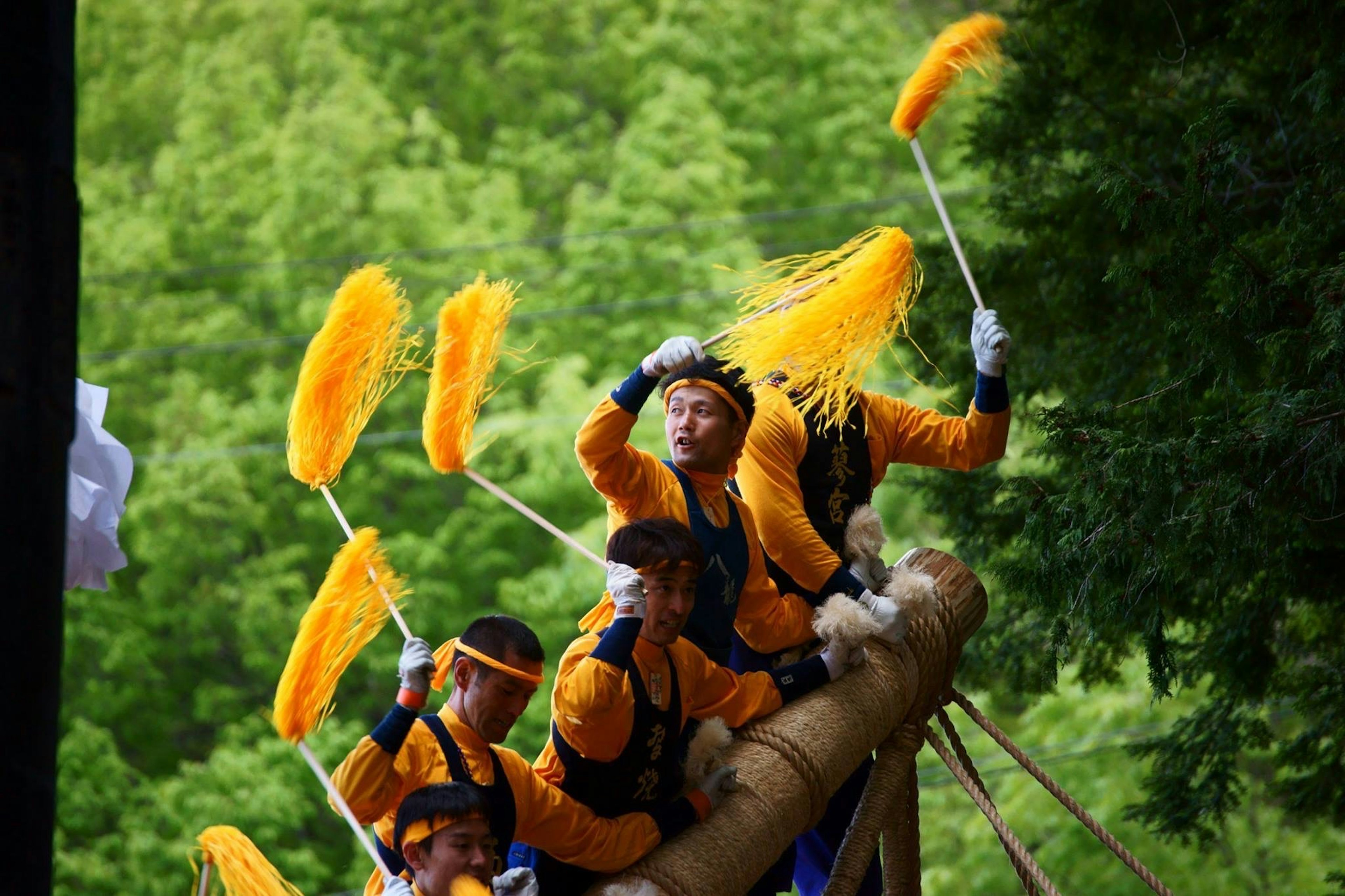 Participants in a festival wearing orange outfits dancing on a boat with yellow feather wands against a green mountain backdrop