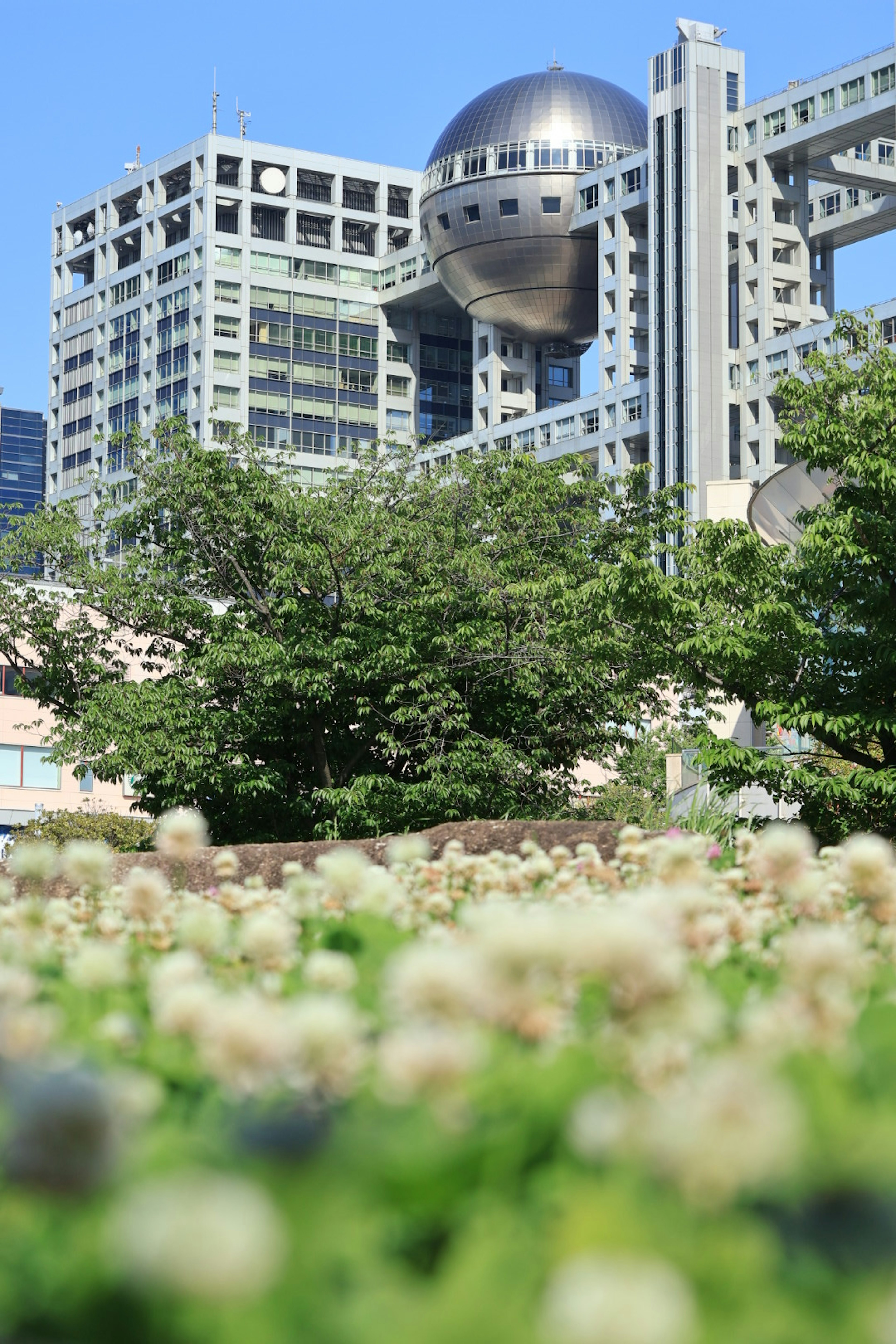 Pemandangan gedung Fuji Television di bawah langit biru dengan taman hijau di latar depan